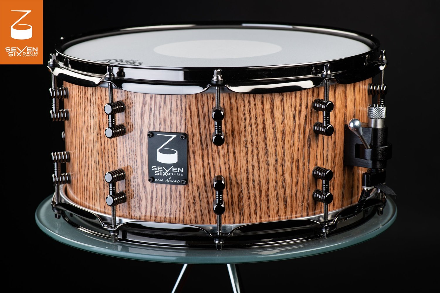 Handmade snare drum featuring a natural oak wood shell with black nickel hoops, dual lugs, and a side throw-off.