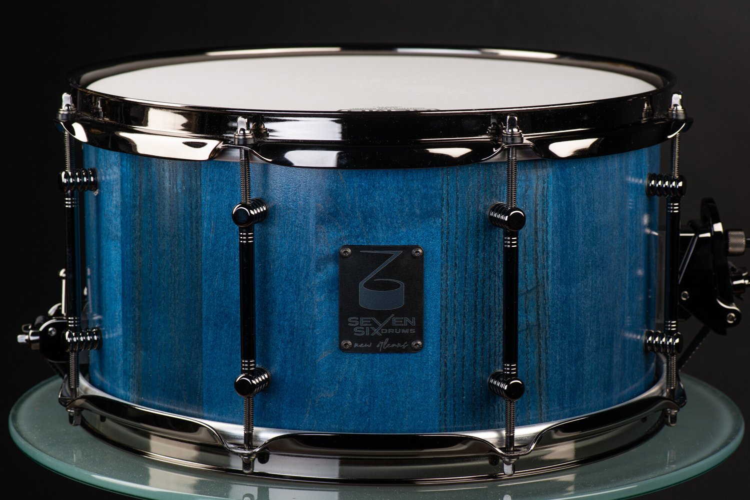 Snare drum featuring a blue-stained wood grain shell, black nickel hoops, and black tube lugs.