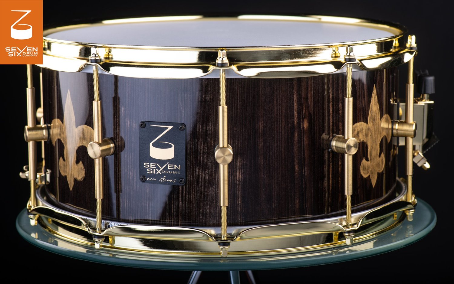 Glossy dark wood snare drum featuring gold fleur-de-lis motifs, polished brass hoops, and brass tube lugs.