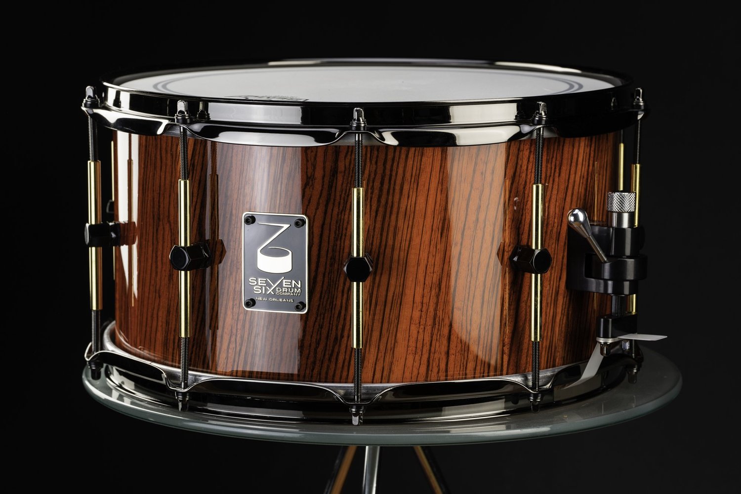 Handmade snare drum with a glossy reddish-brown wood shell, dark chrome hoops, and brass tube lugs.