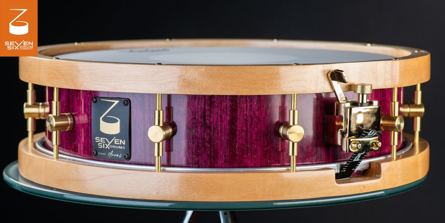 Handmade snare drum with a glossy purple wood shell, natural wood hoops, brass lugs, and a gold throw-off.