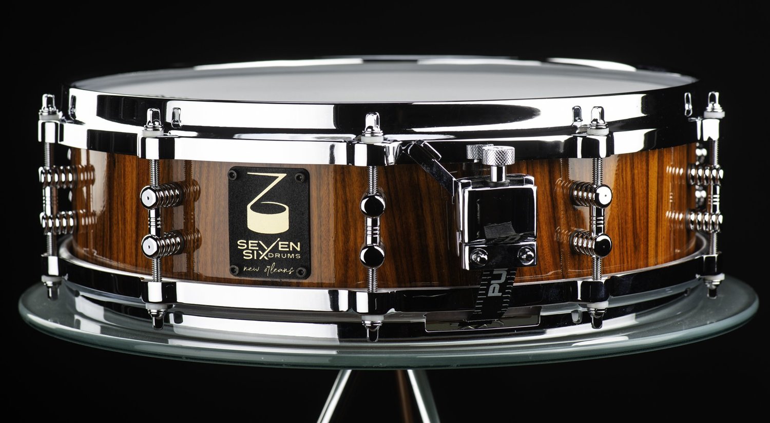 Handmade snare drum featuring a glossy striped wood shell, polished chrome hoops, dual-point lugs, and a throw-off.