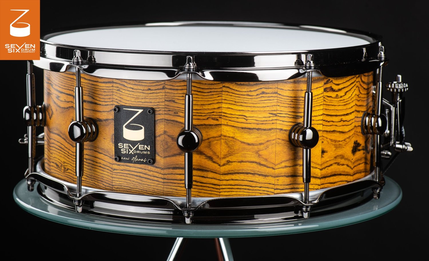 Handcrafted snare drum with a natural striped segmented wood shell, black nickel hoops, and tube lugs.