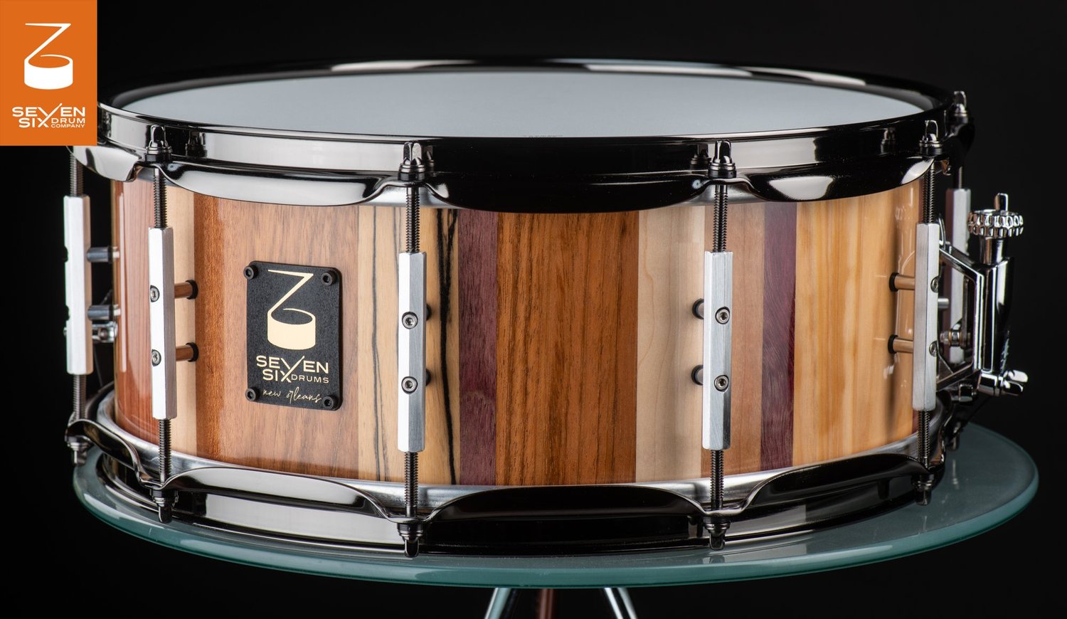 Handmade snare drum featuring a striped mixed-wood shell with a natural finish, brushed silver lugs, and black nickel hoops.