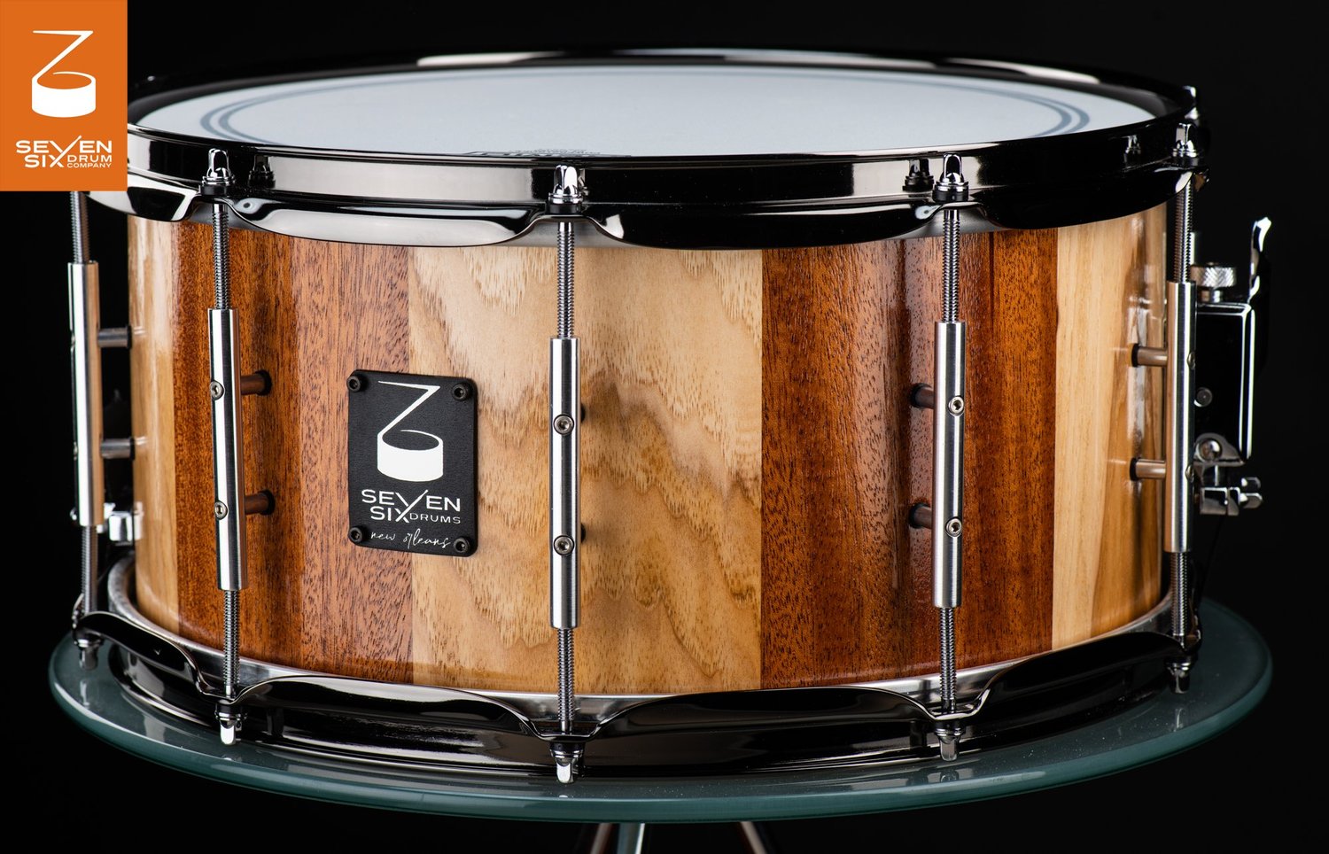 Handmade snare drum featuring a glossy striped wood shell, polished chrome tube lugs, and black nickel hoops.