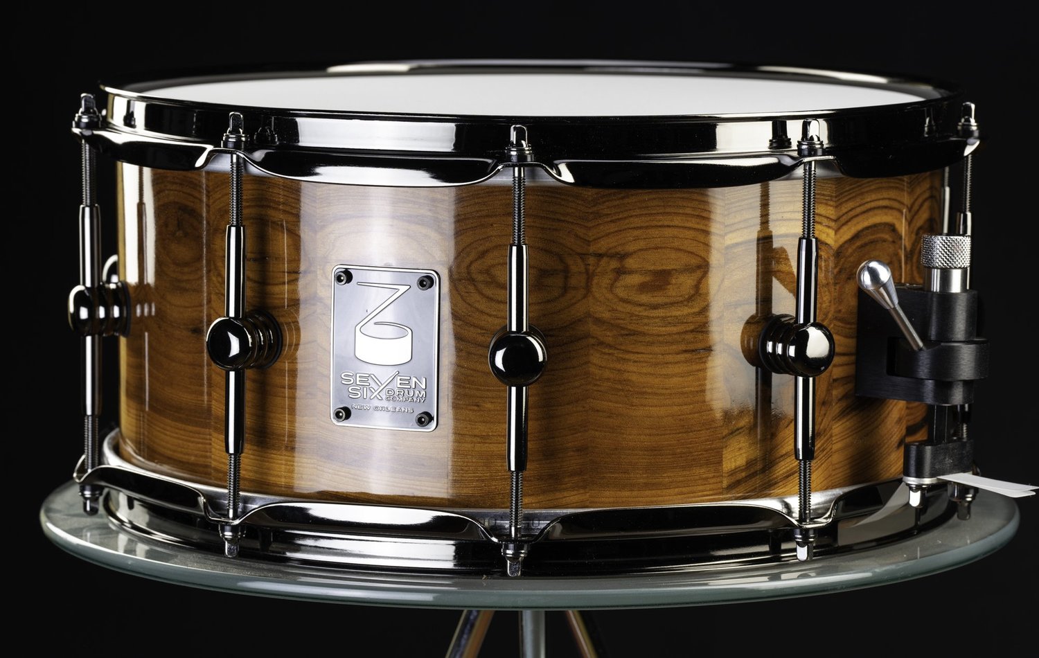 Handmade snare drum with a glossy wood grain shell, black nickel hoops, center lugs, and a snare throw-off.