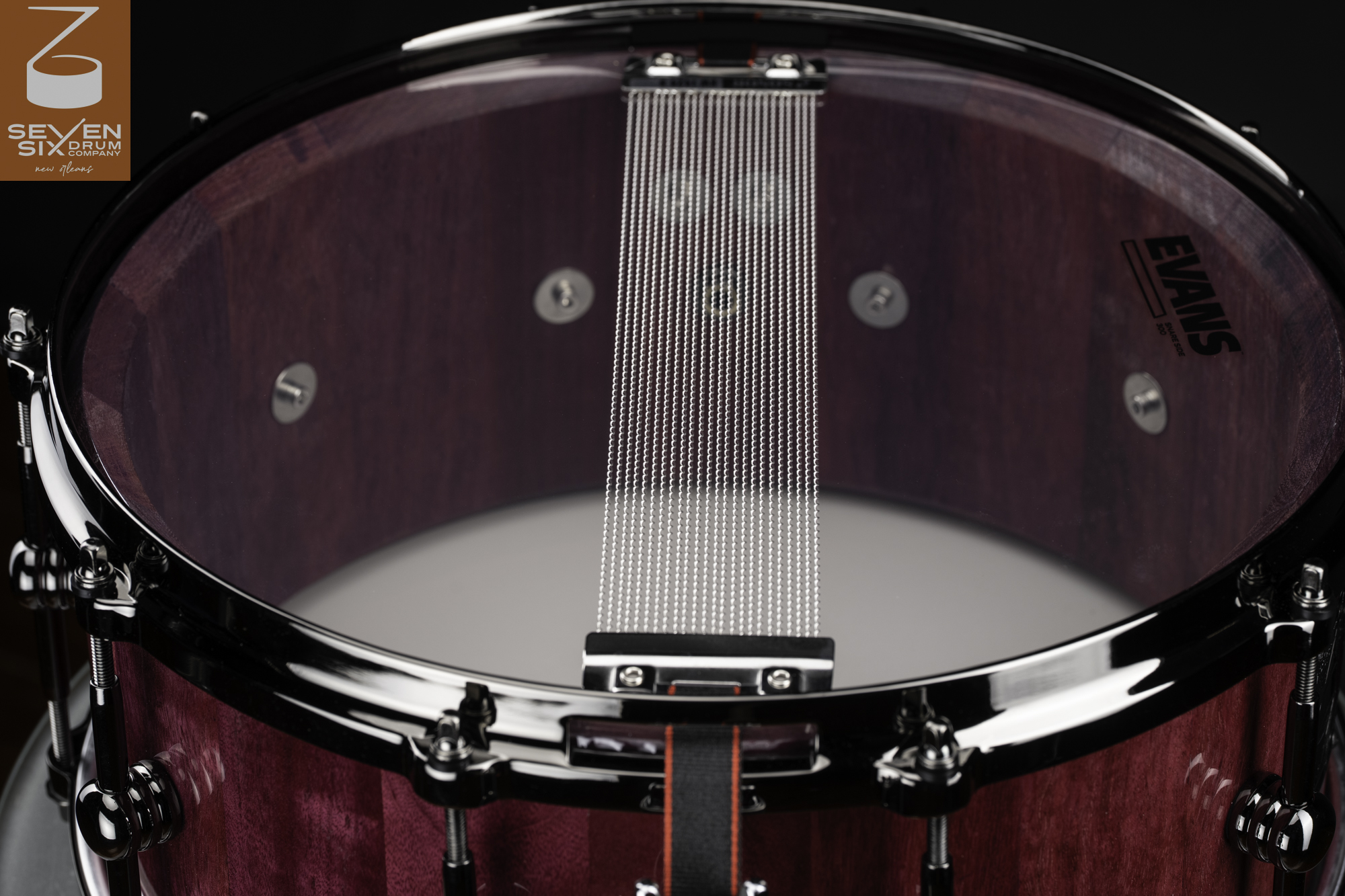 Snare-side view of a purpleheart wood drum with wire snares, a clear resonant head, and gloss black hardware.