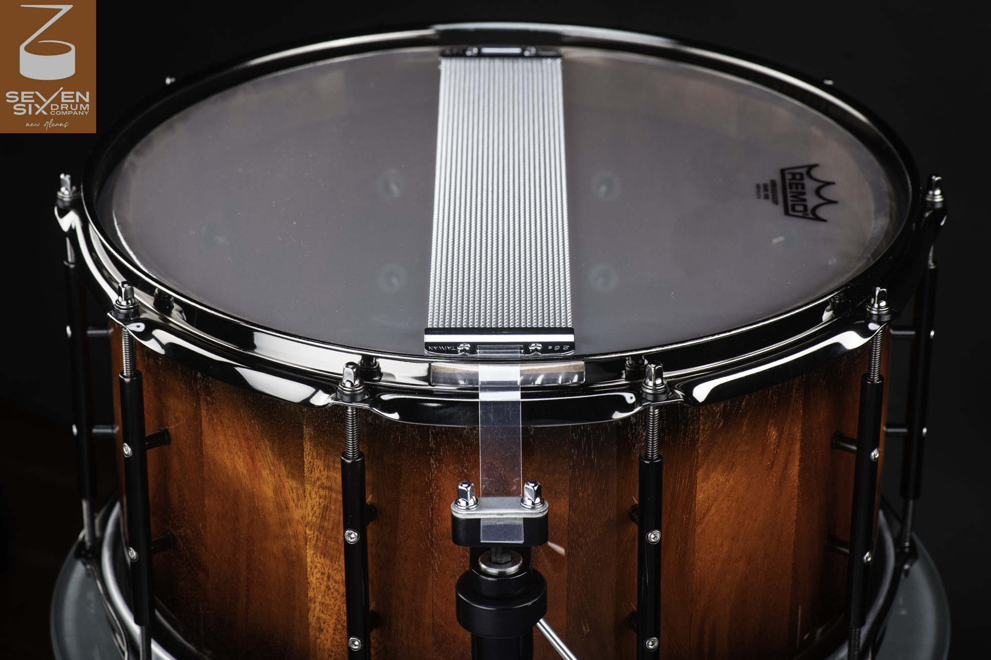 Bottom view of a handmade reddish wood snare drum with chrome snare wires, black hardware, and a clear resonant head.