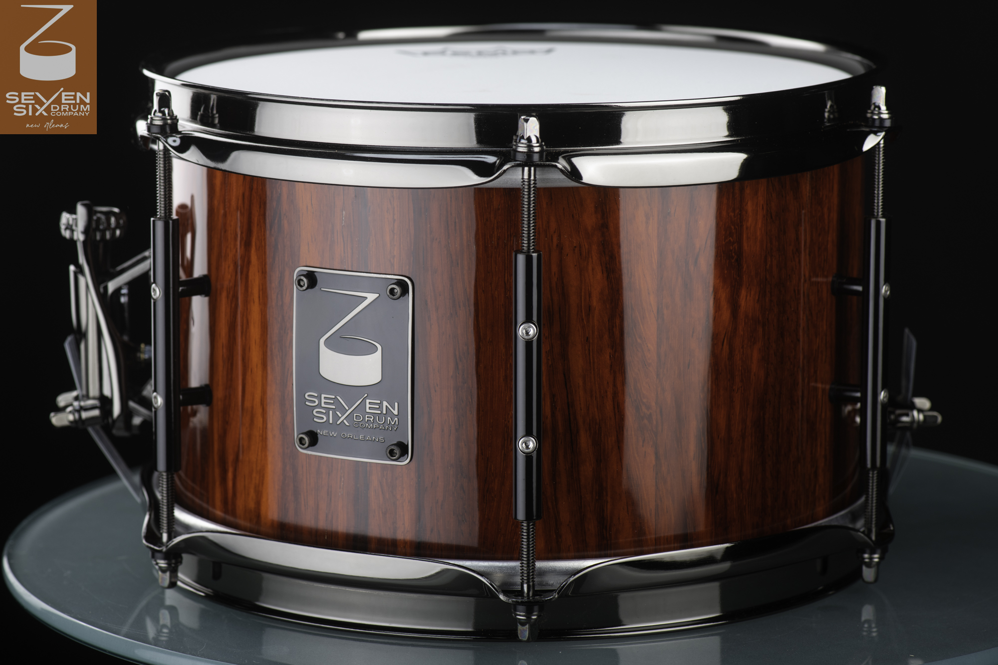 Snare drum featuring a glossy reddish-brown wood shell, black nickel hoops, and matching black tube lugs.