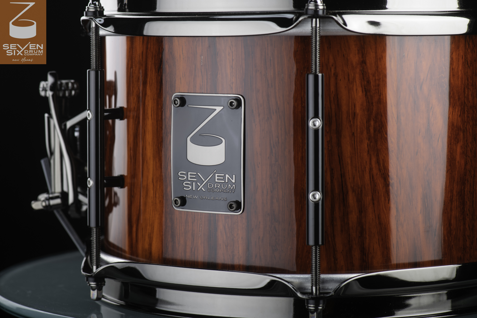 Glossy reddish-brown wood snare drum featuring black tube lugs, black chrome hoops, and a Seven Six Drum Company badge.
