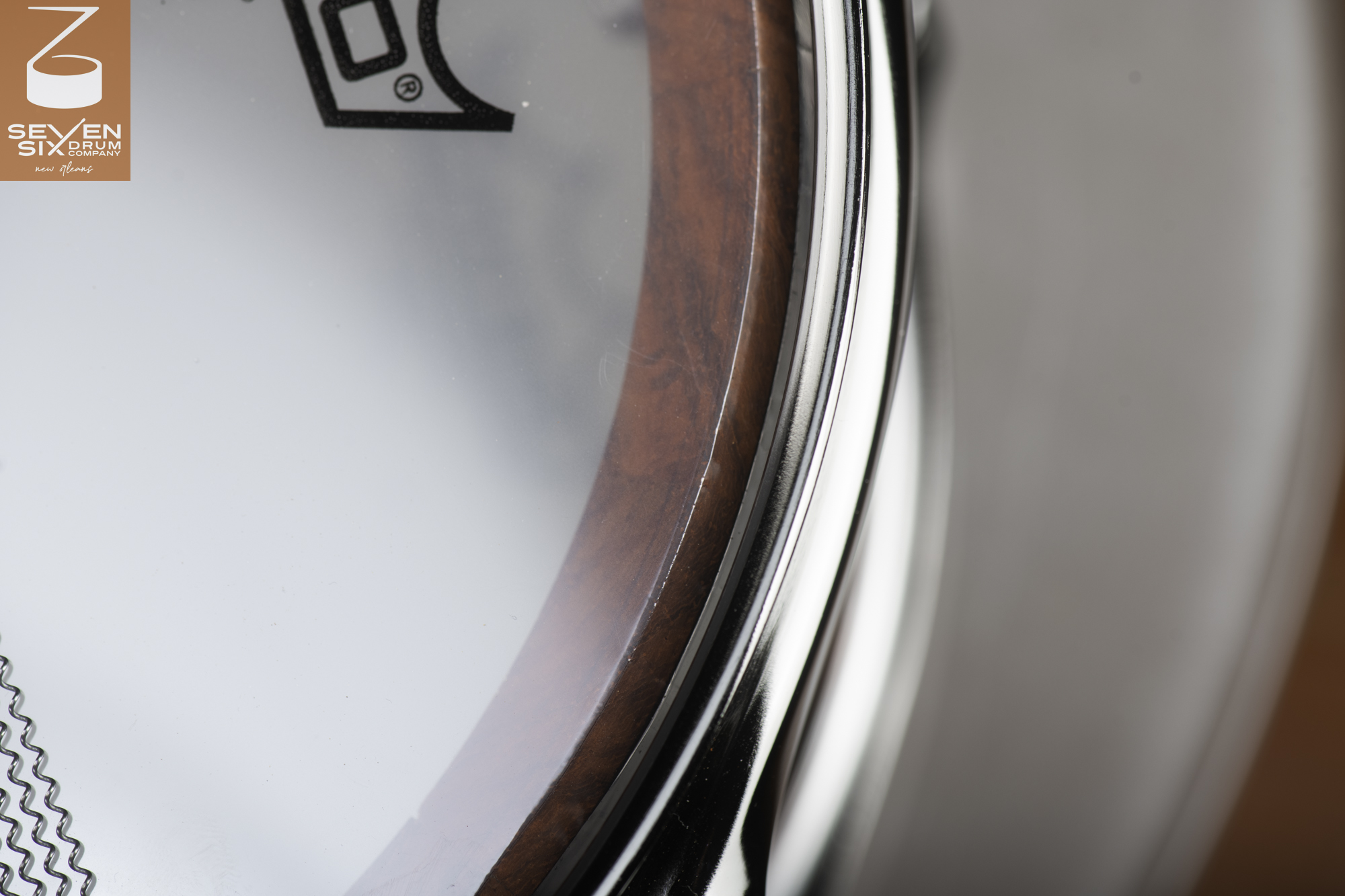 Close-up of a snare drum showing a reddish-brown wood shell, polished chrome hoop, clear drumhead, and snare wires.