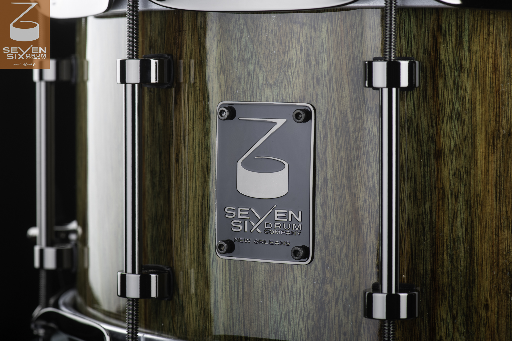 Glossy wood grain snare drum shell with chrome tube lugs and a Seven Six Drum Company metal badge.