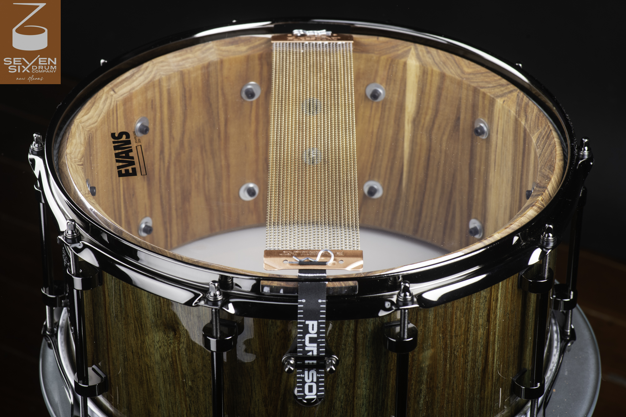 Bottom of a glossy natural wood snare drum featuring black hardware, a clear Evans head, and copper snare wires.