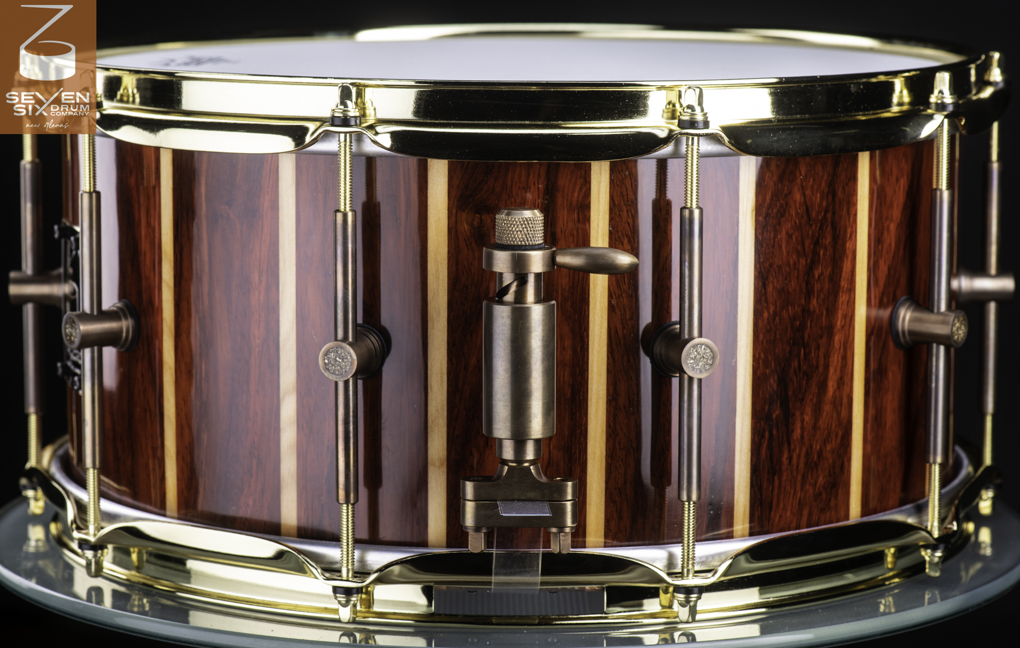 Handmade snare drum featuring a glossy striped wood shell, polished brass hoops, and an antique bronze throw-off.