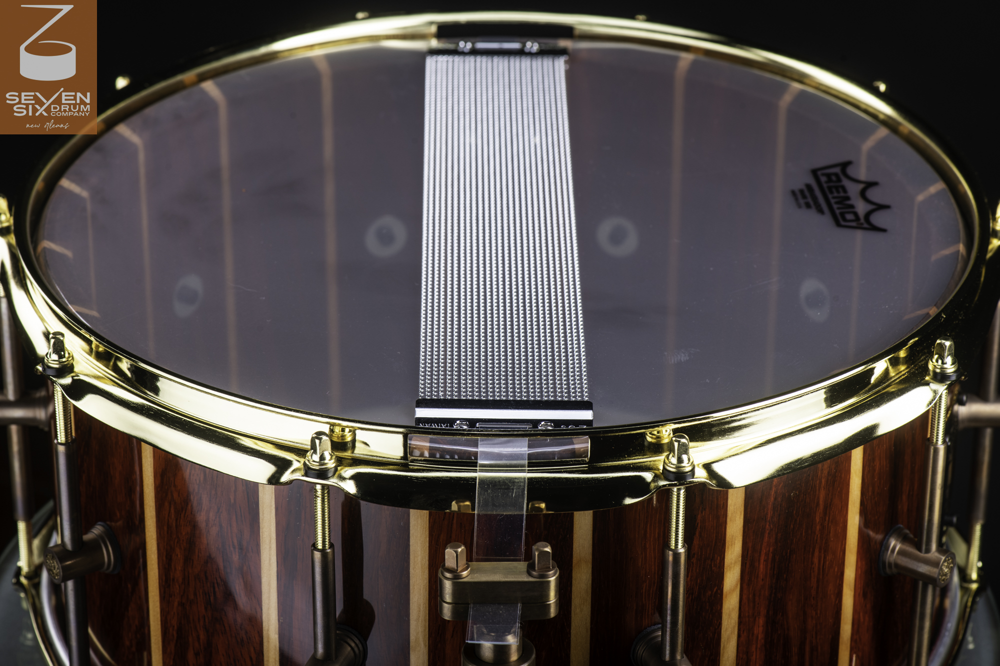 Glossy striped wood snare drum with a clear resonant head, steel snare wires, bronze lugs, and a polished brass hoop.