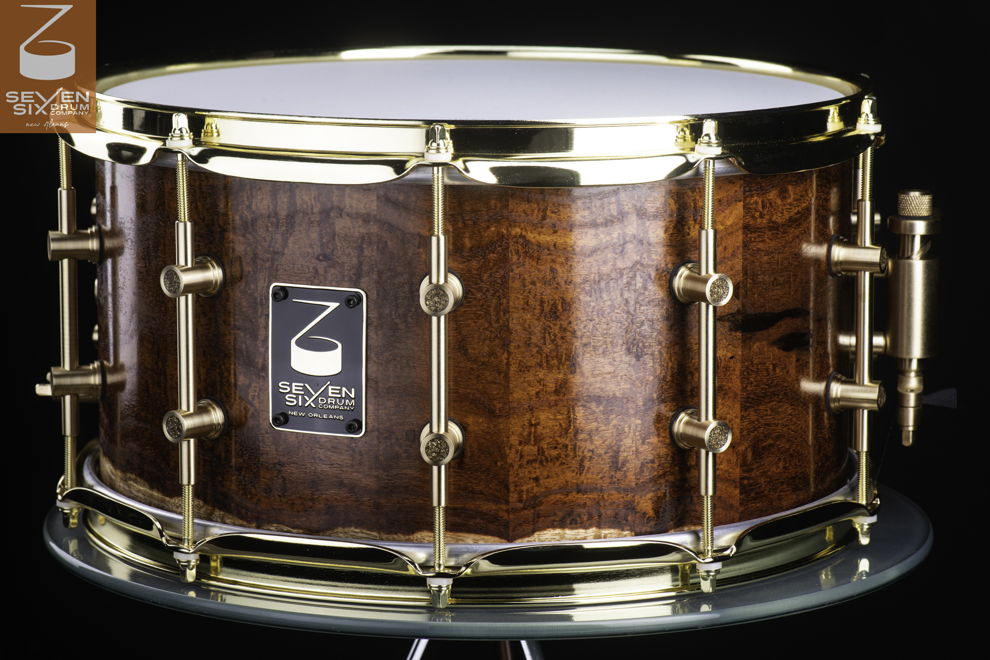 Handmade snare drum with a glossy figured brown wood shell, polished brass hoops, brass tube lugs, and a matching throw-off.