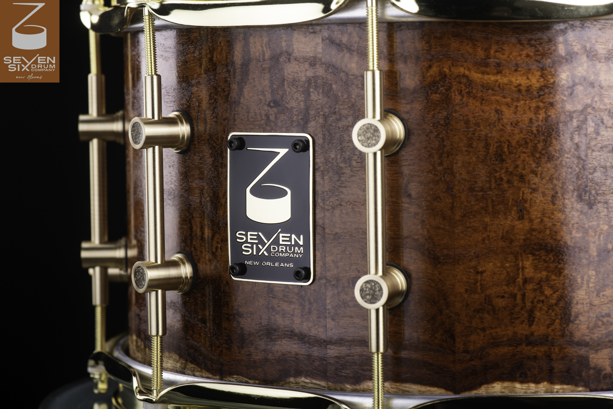 Glossy brown figured wood snare drum with brushed brass tube lugs, hoops, and a black branded badge.