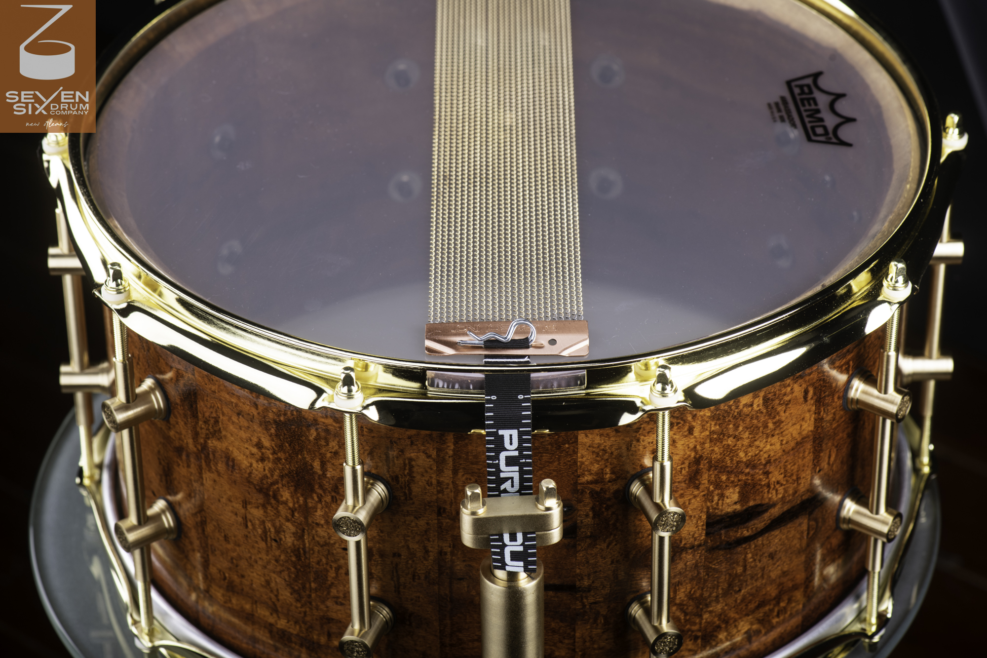 Handmade snare drum with a glossy figured wood shell, brass hoops, tube lugs, and snare wires over a clear head.