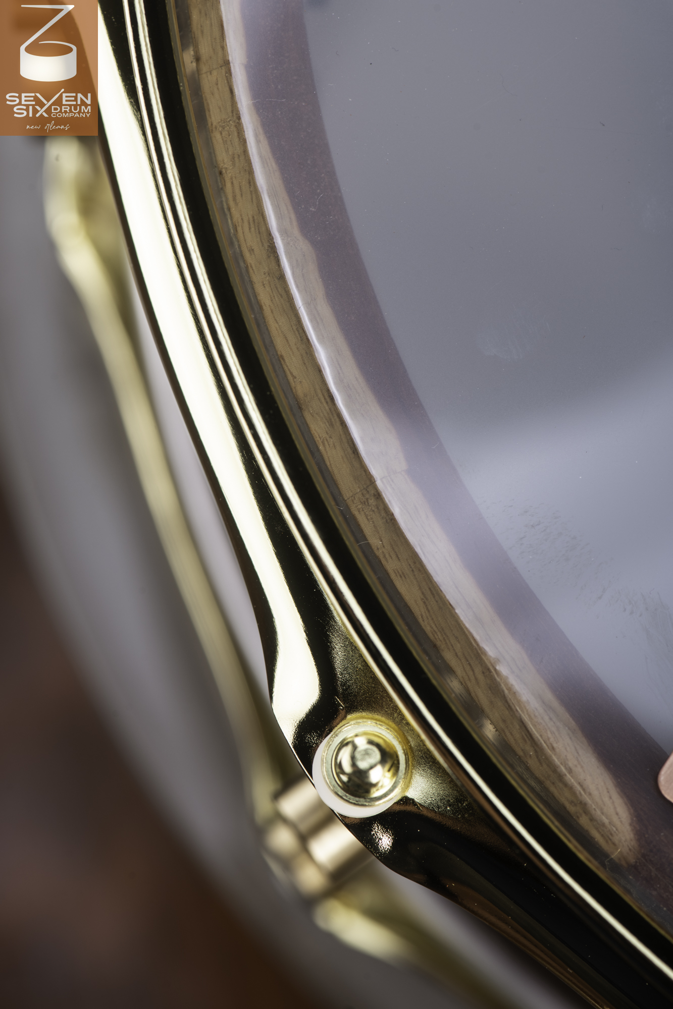 Close-up of a natural wood snare drum with a clear head, showing a polished gold hoop and tension rod hardware.