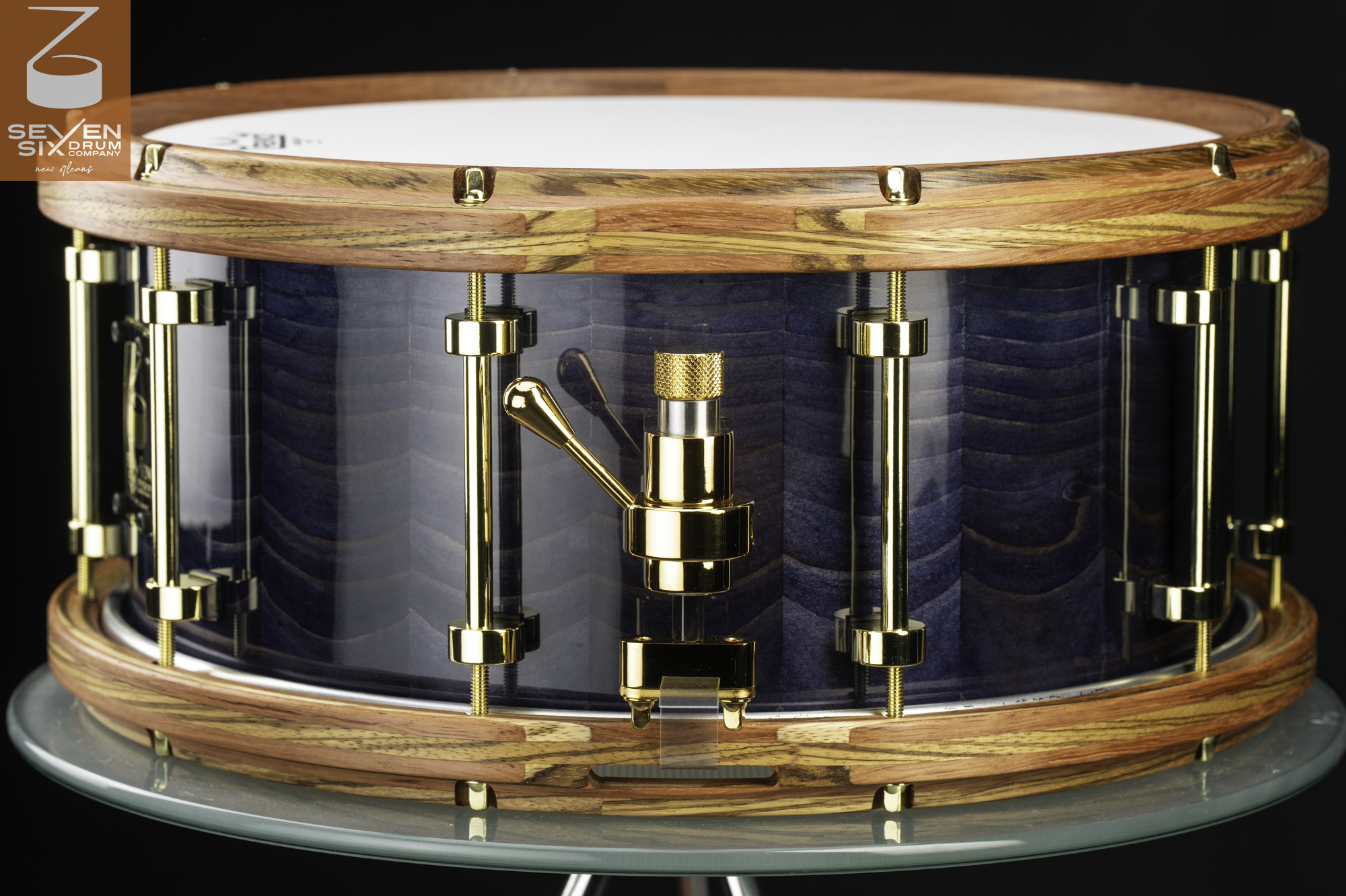 Handmade snare drum with a glossy blue figured wood shell, segmented wooden hoops, and polished gold hardware.