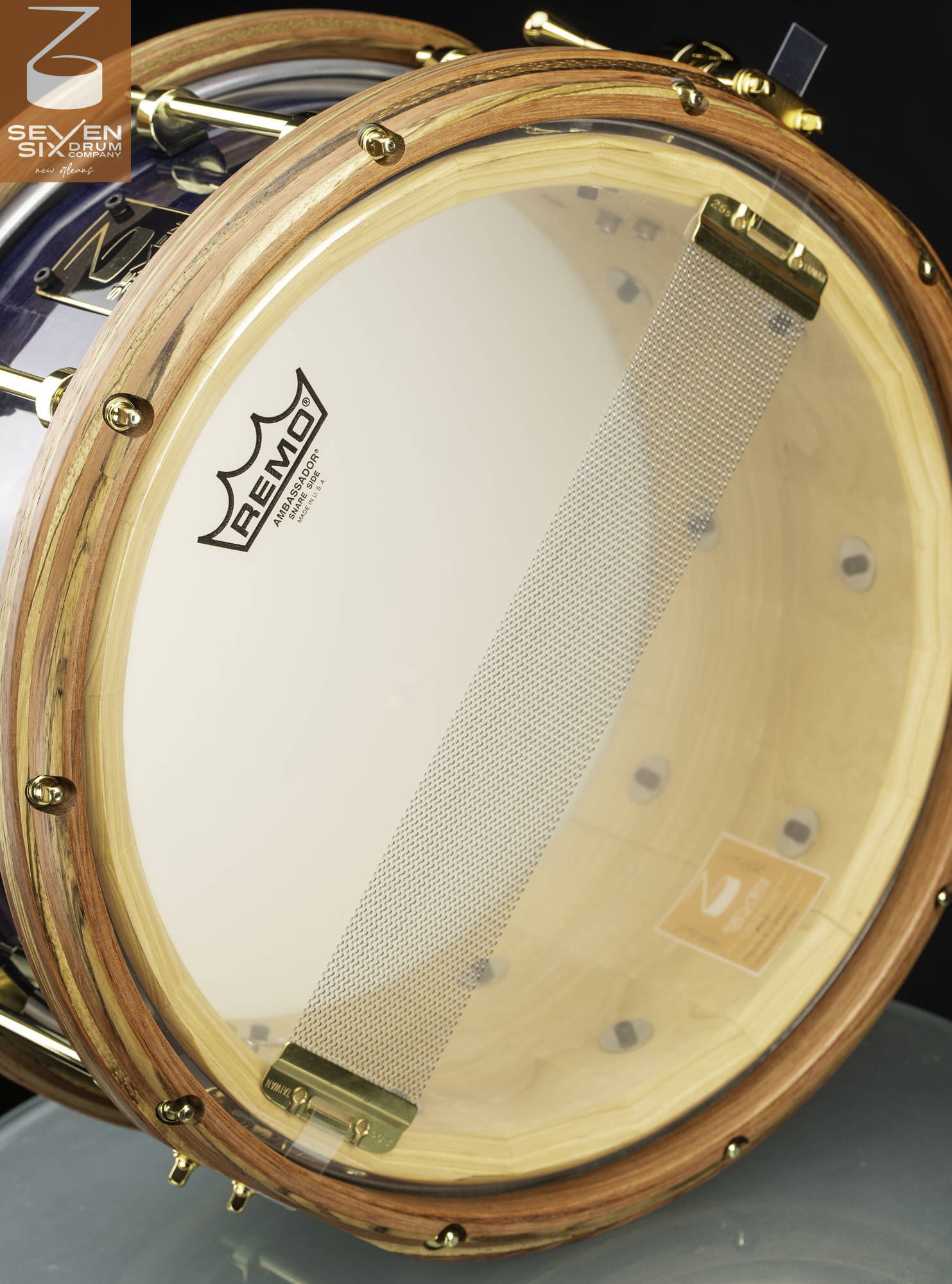 Bottom view of a custom snare drum featuring natural wood hoops, polished brass hardware, and snare wires on a clear head.