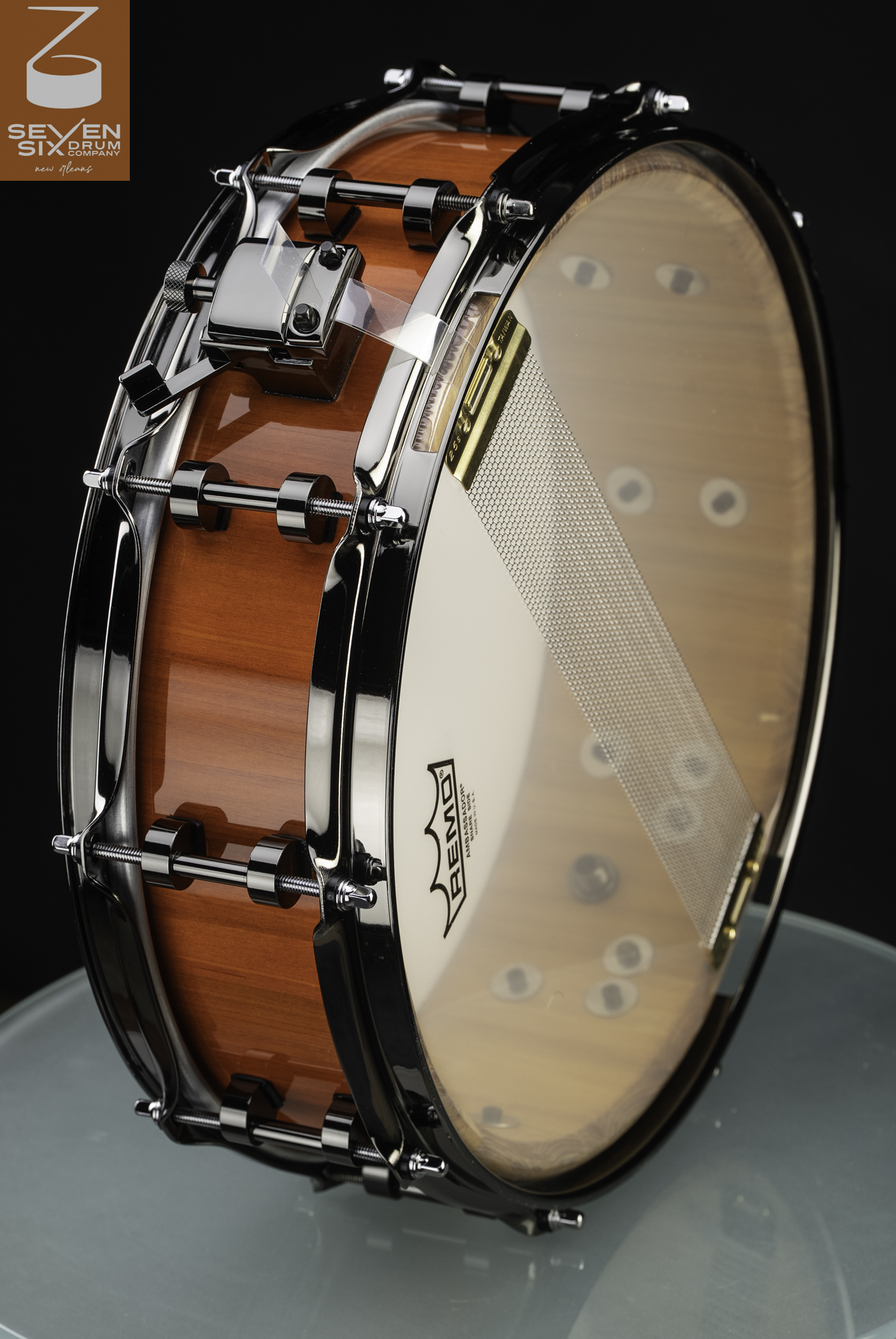 Handmade snare drum with a glossy amber wood shell, black nickel hardware, and snare wires on a clear head.