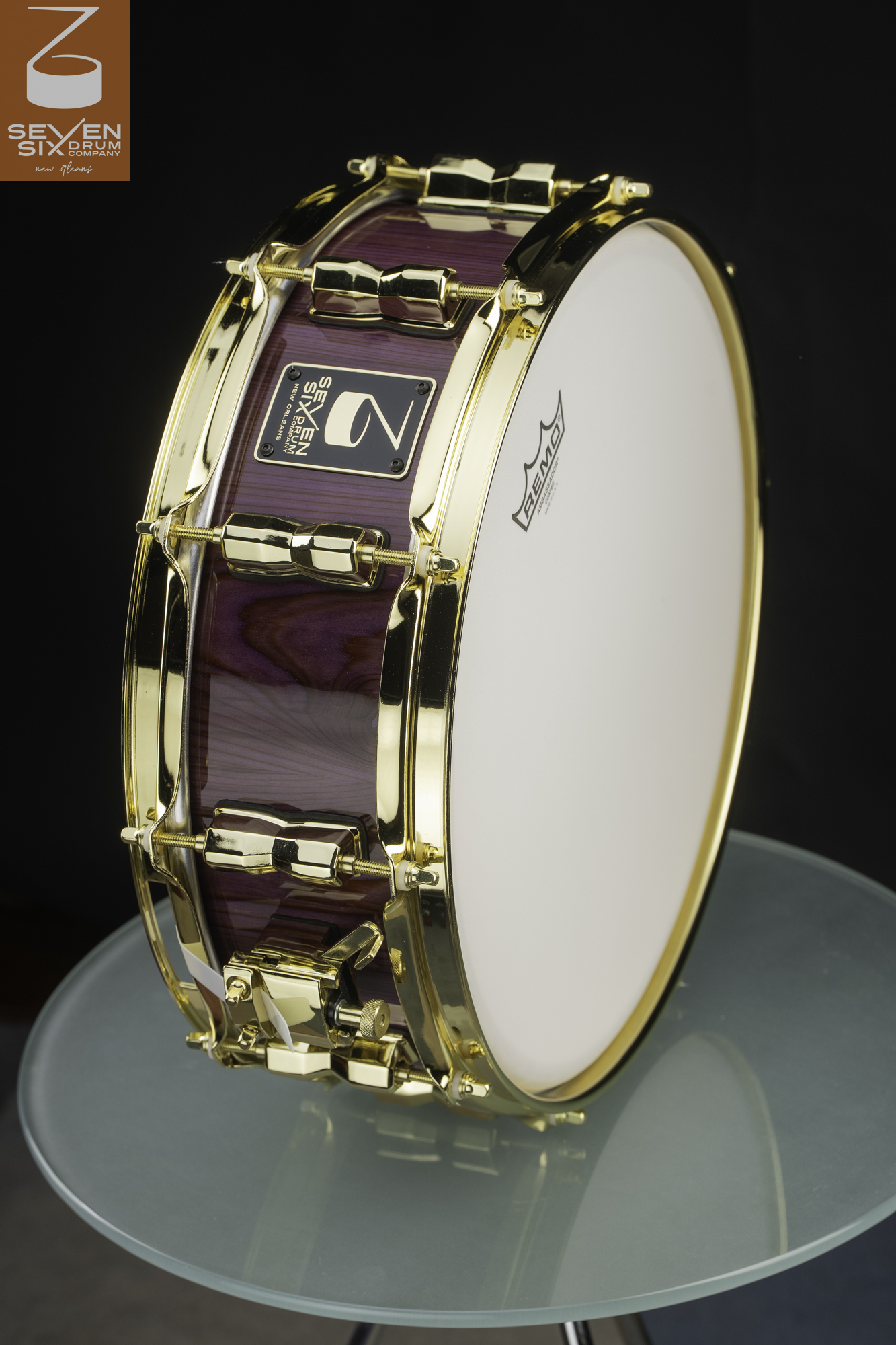 Handmade snare drum featuring a high-gloss purple wood shell with polished gold hoops, lugs, and throw-off.