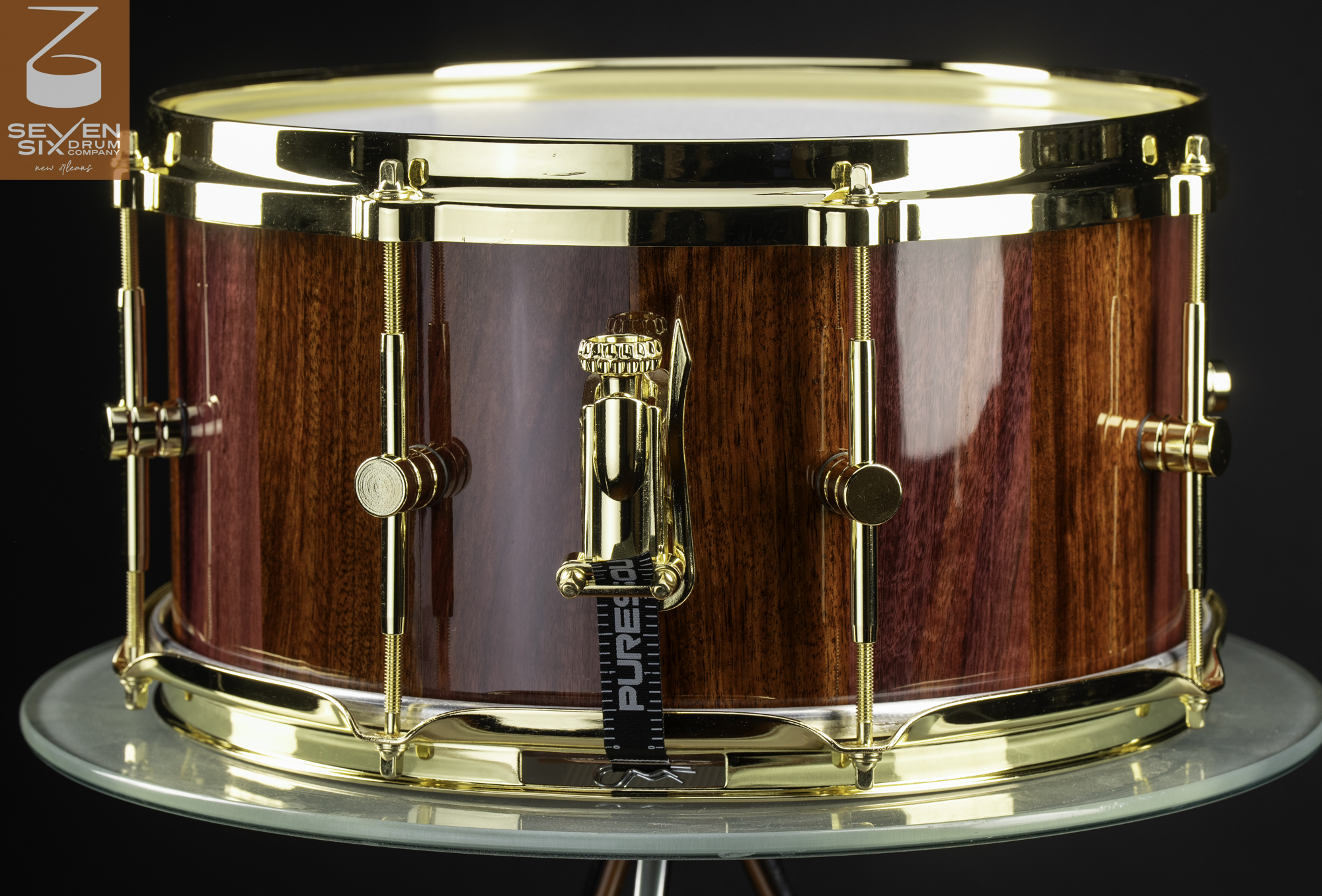 Handmade snare drum featuring a glossy reddish wood shell with polished gold hoops, lugs, and throw-off.