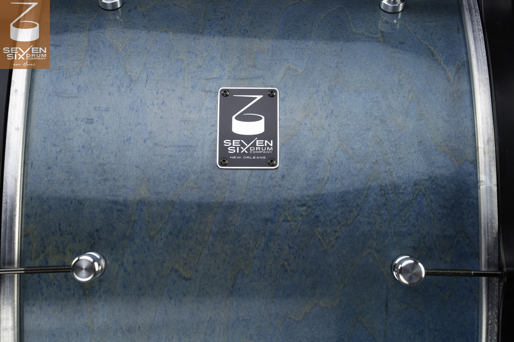 Glossy blue-stained figured wood drum shell with chrome hardware and a black Seven Six Drum Company badge.