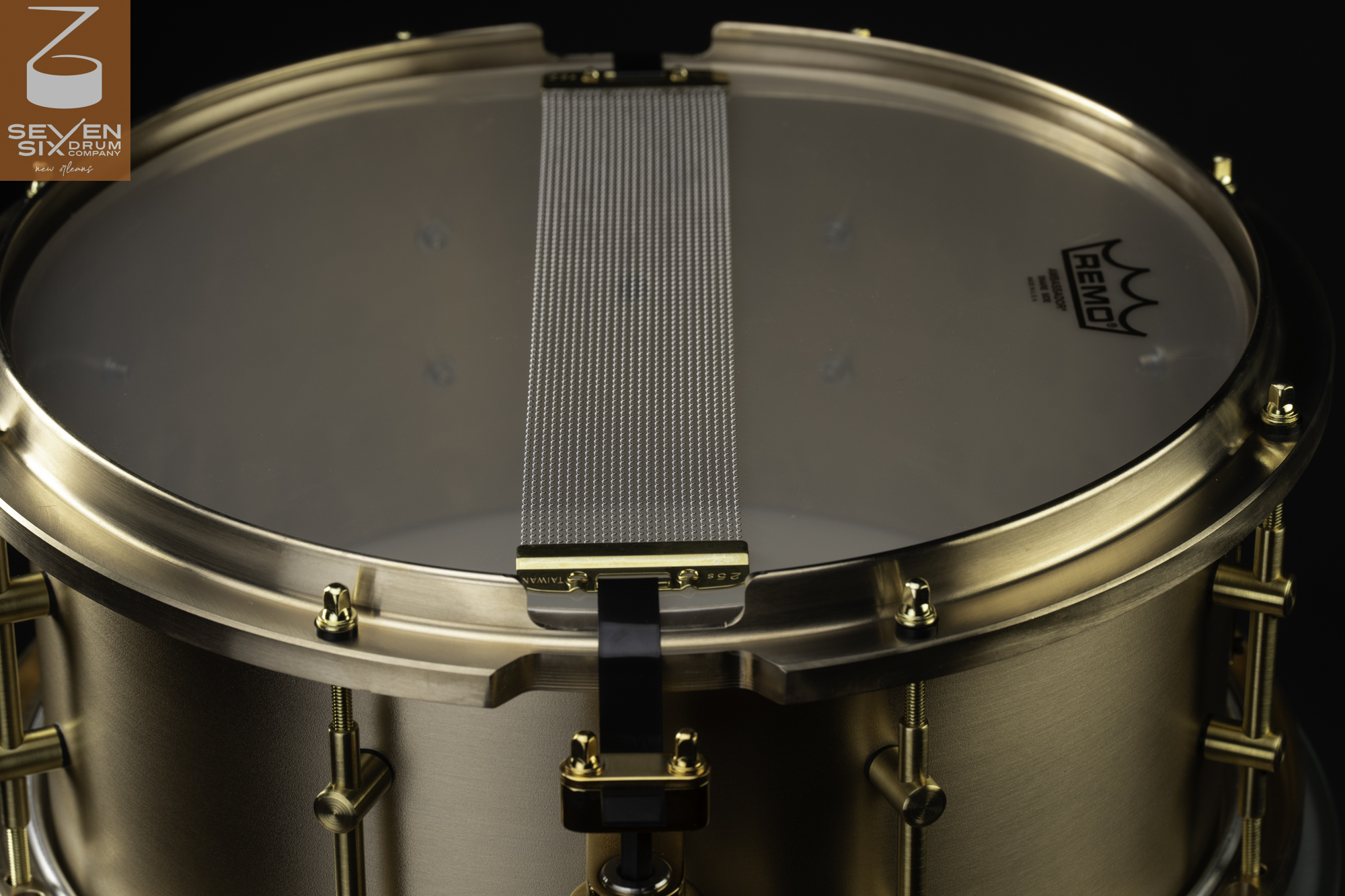 Underside of a brushed brass snare drum featuring wire snares over a clear head, a contoured metal hoop, and tube lugs.