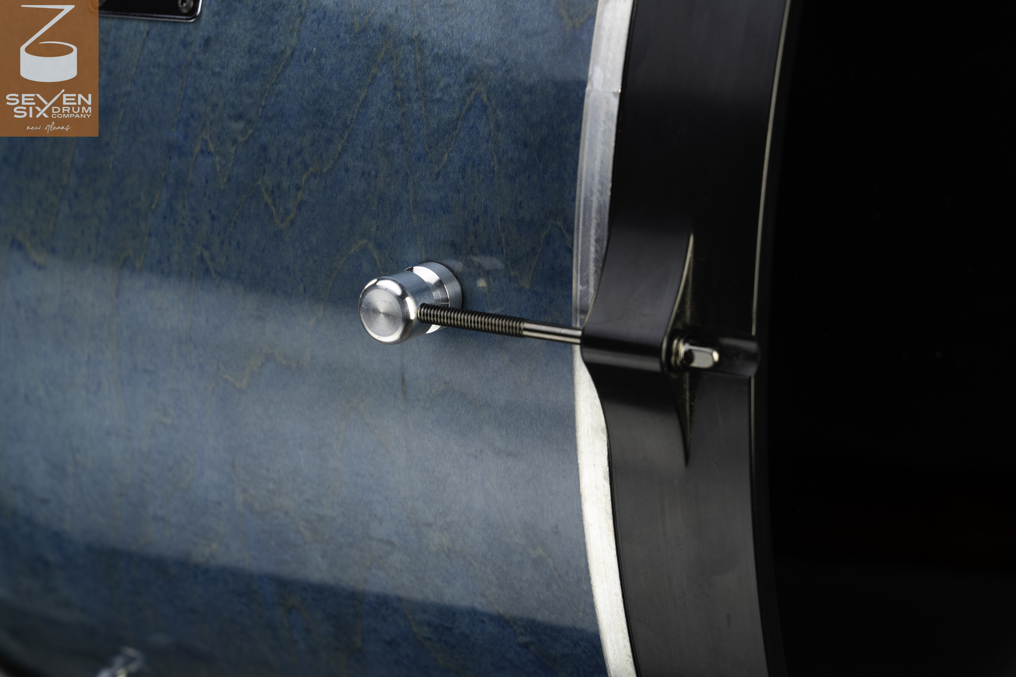 Close-up of a blue figured wood drum shell with a silver round lug, threaded tension rod, and black contoured hoop.