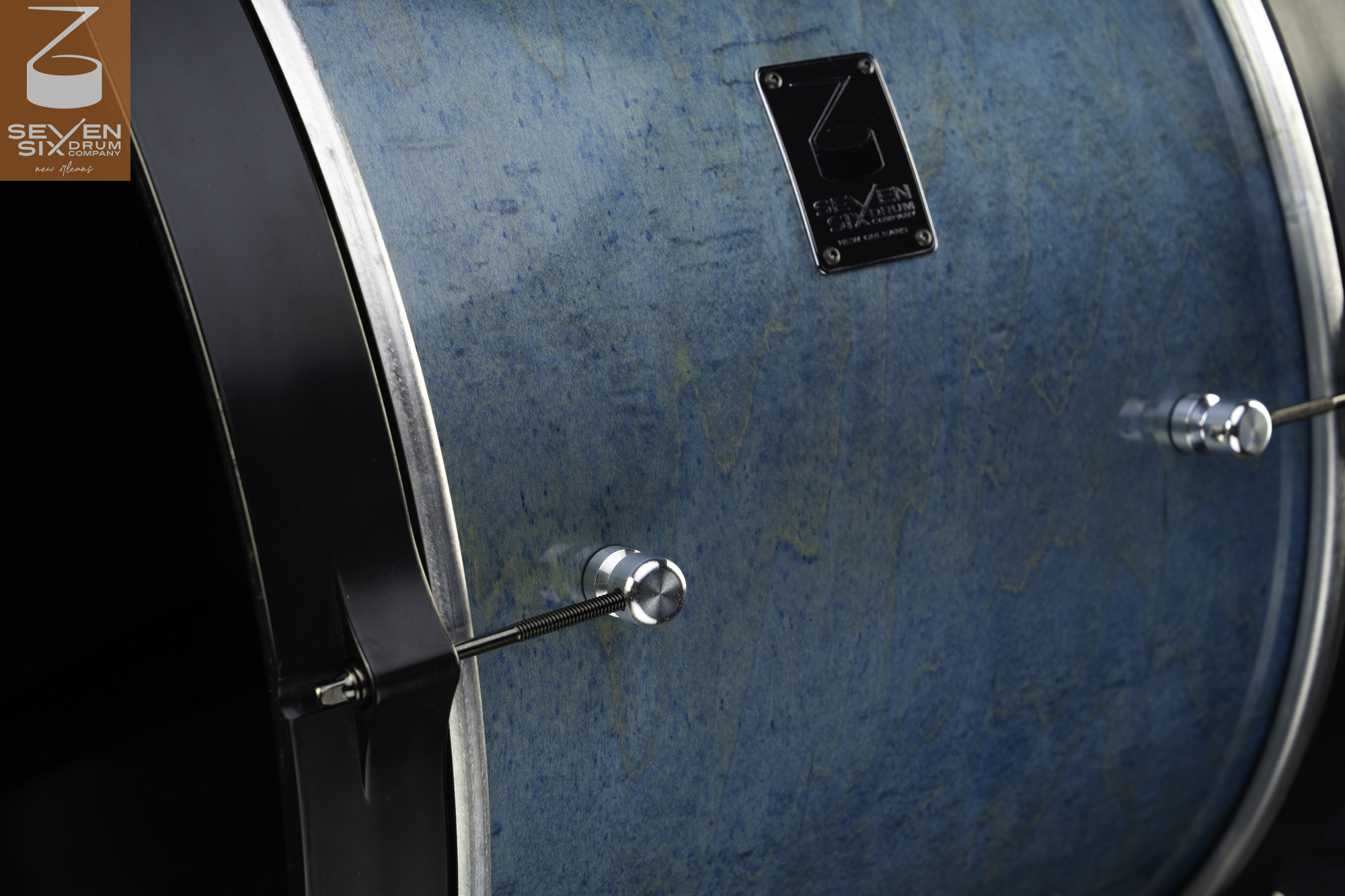 Blue-stained figured wood drum shell featuring round chrome lugs, black tension rods, and a black wooden hoop.