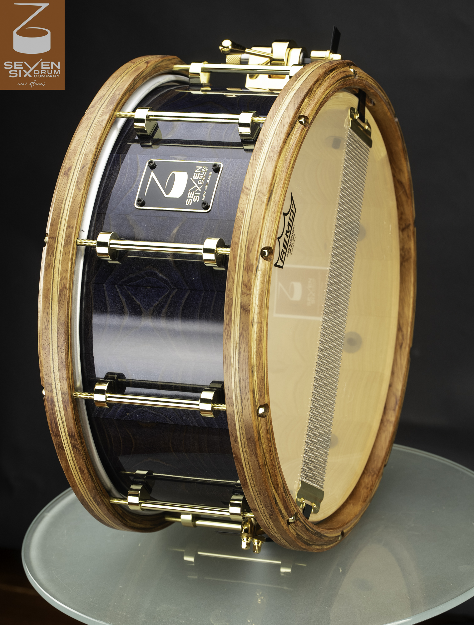 Handmade snare drum with a glossy dark blue wood shell, thick natural wood hoops, and polished gold tube lugs.