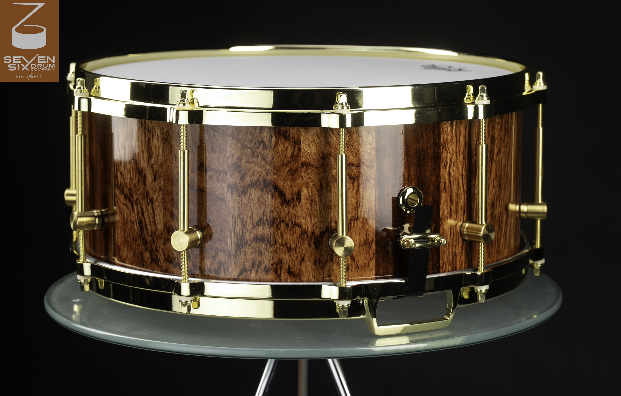 Handmade snare drum with a high-gloss figured wood shell, polished gold hoops, and brass tube lugs.