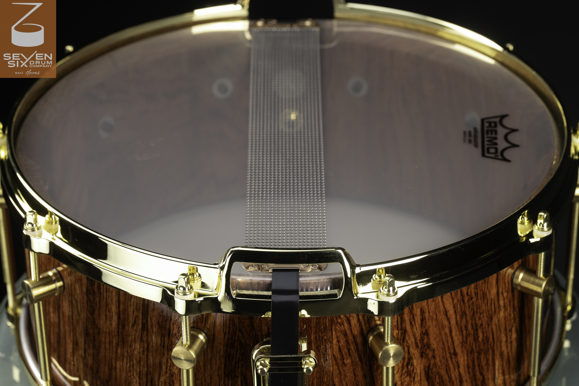 Bottom of a polished natural wood snare drum featuring gold-plated hoops, brass lugs, and steel snare wires.
