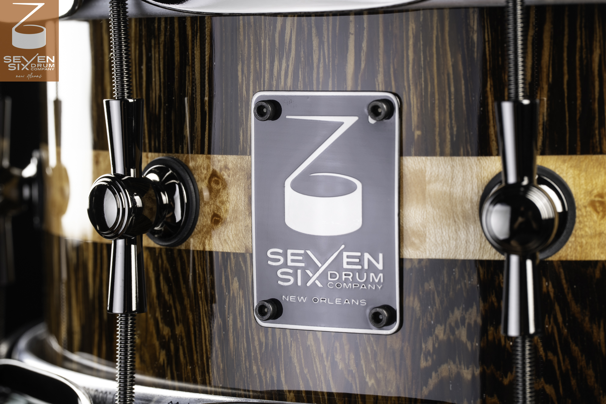 Glossy dark wood snare drum featuring a light burl center stripe, black nickel lugs, and a Seven Six Drum Company metal badge.