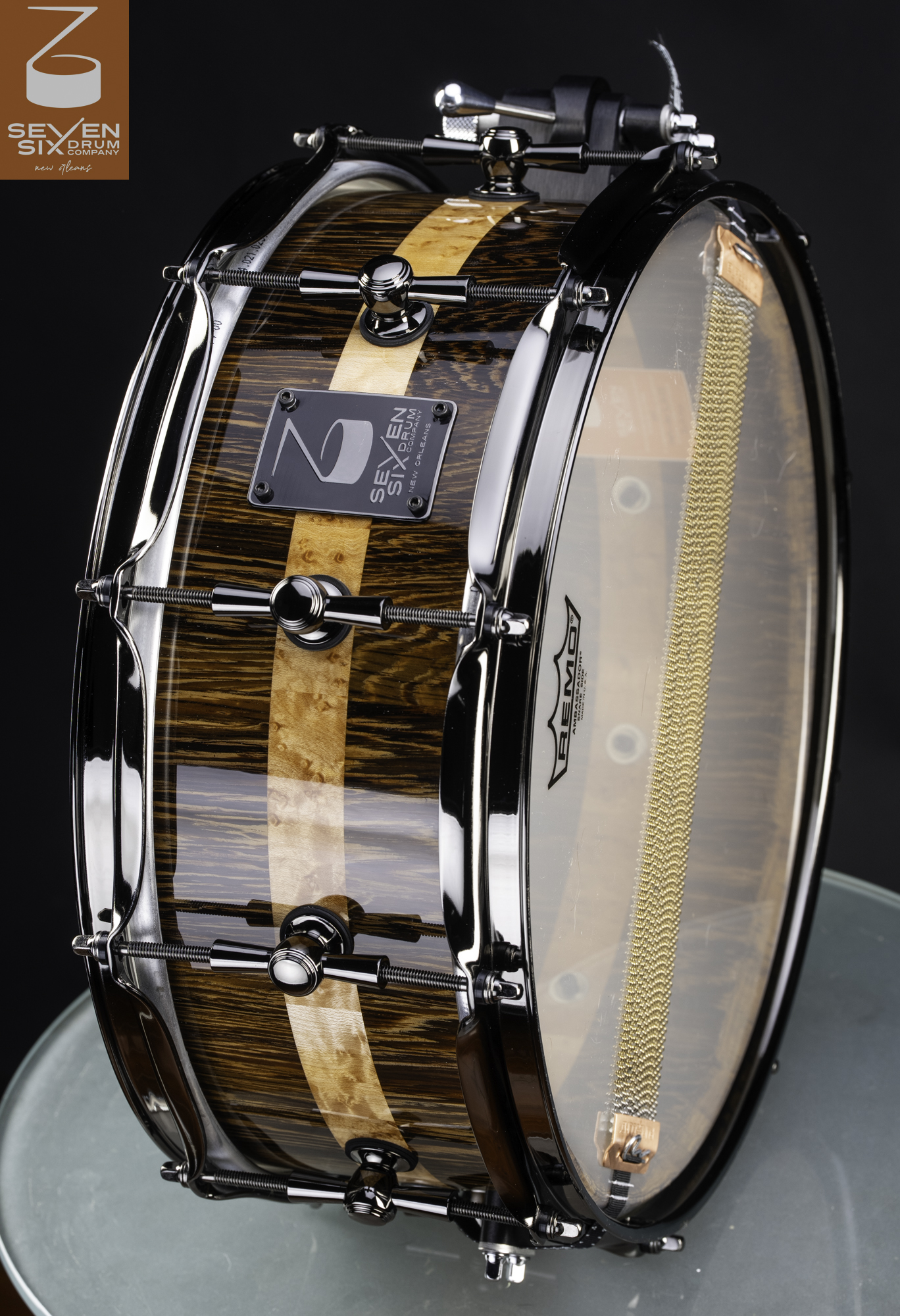 Handmade snare drum with a high-gloss figured wood shell, light burl center stripe, and black nickel hardware.