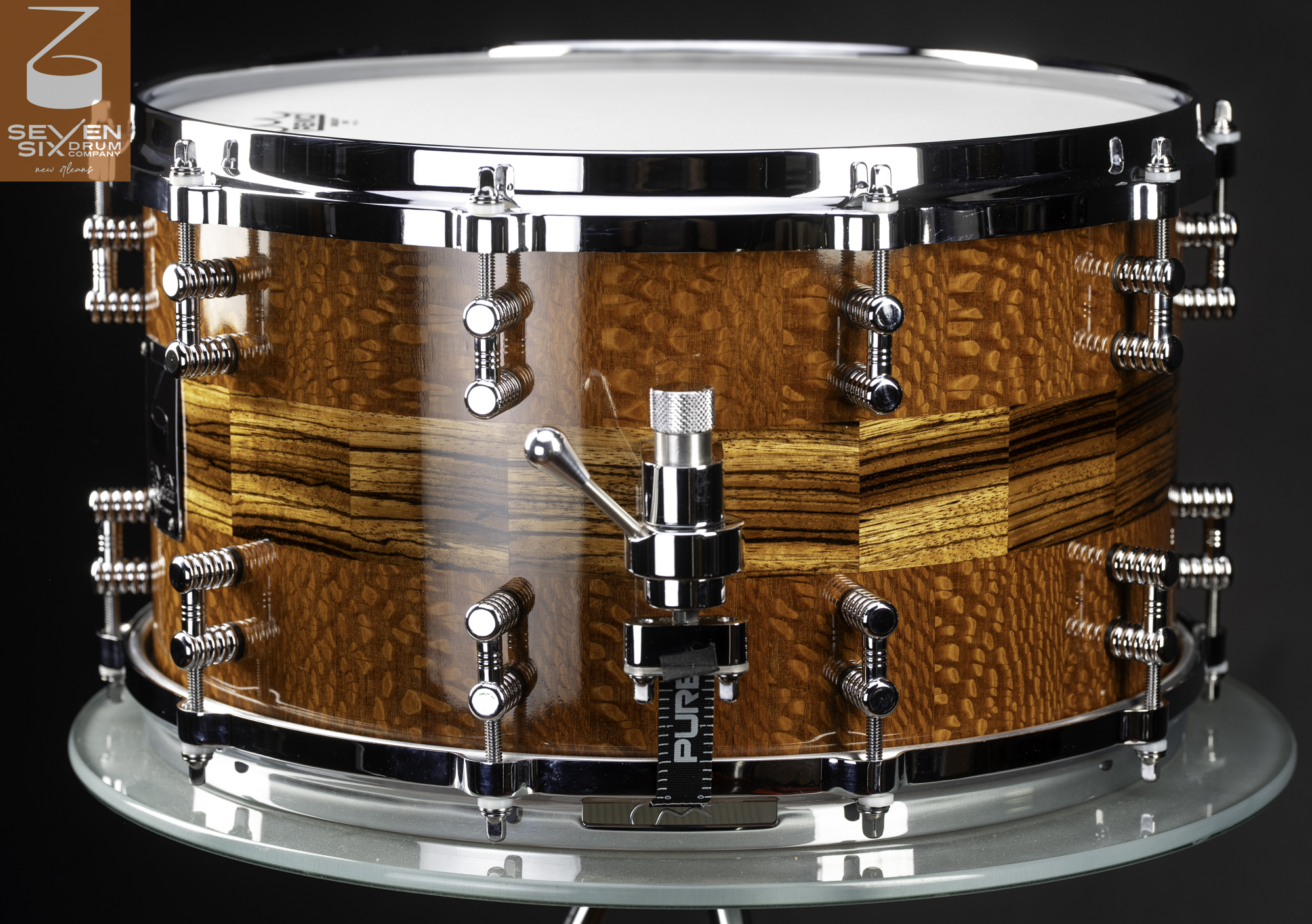 Handmade snare drum in glossy figured wood with a striped inlay, featuring chrome hoops, lugs, and a throw-off strainer.