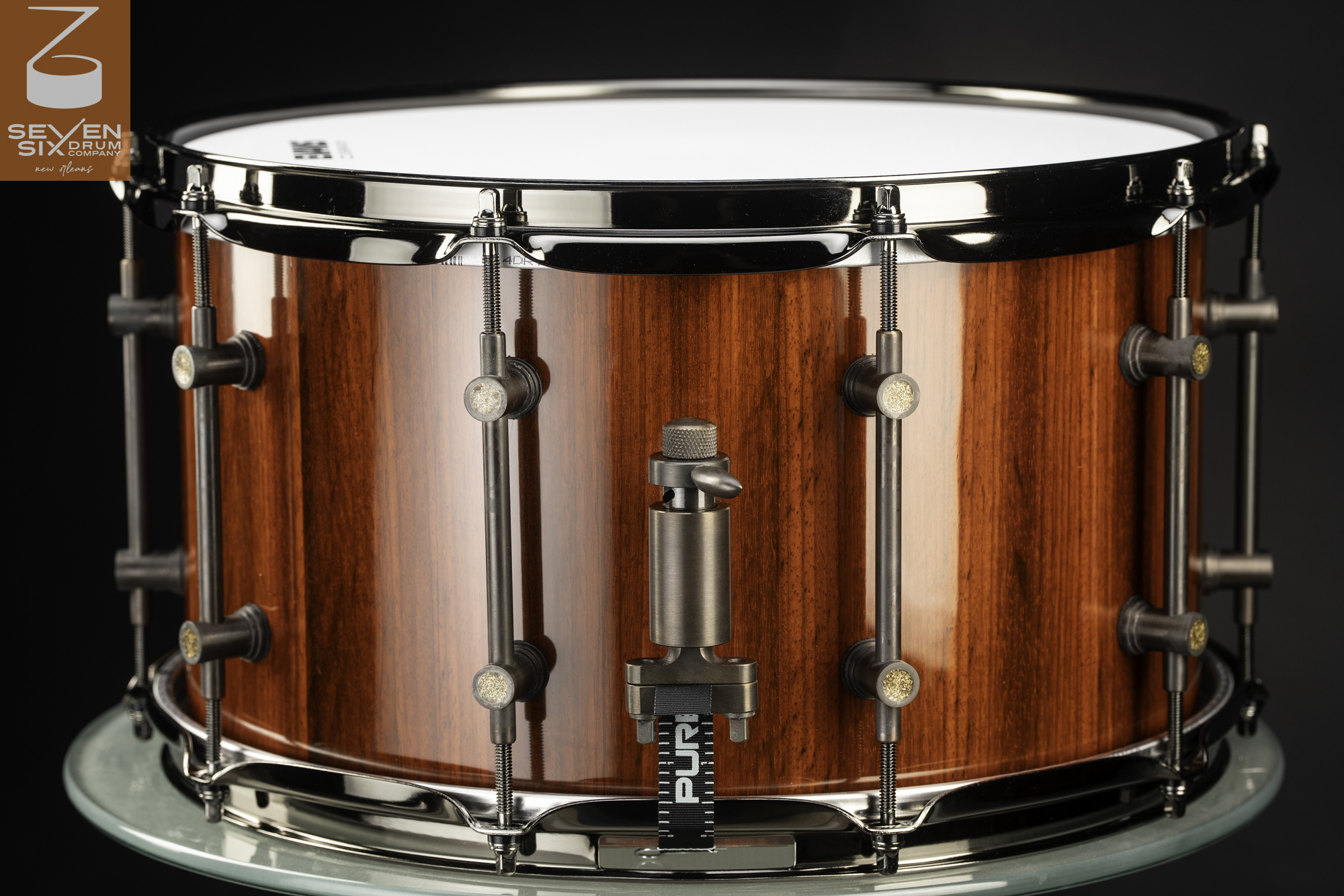 Handmade snare drum with a glossy reddish-brown wood shell, dark nickel hoops, tube lugs, and a snare throw-off.