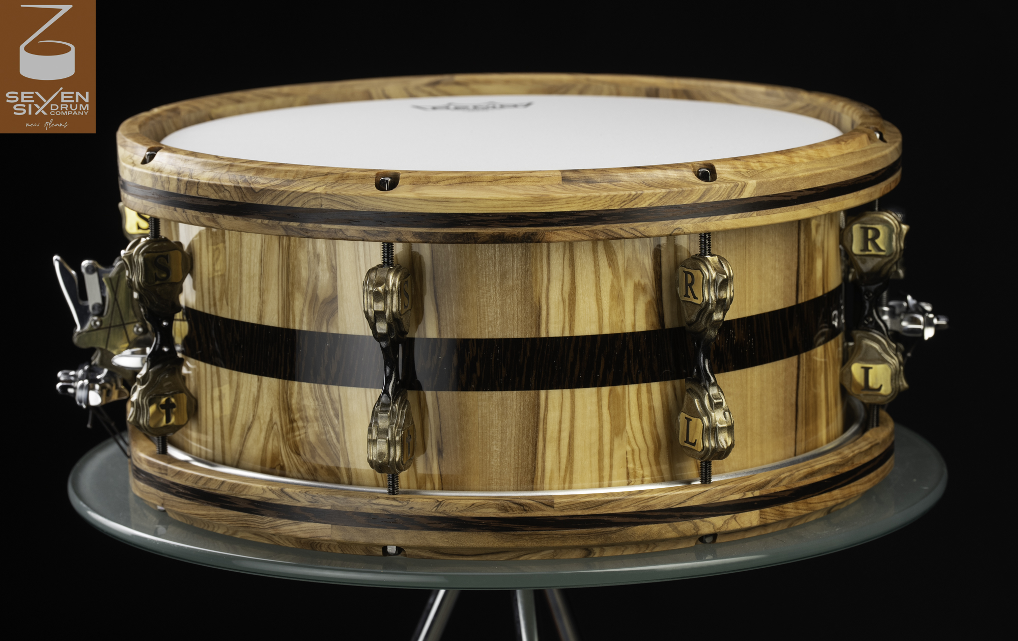 Glossy wood snare drum with a dark horizontal stripe, matching wooden hoops, and antique brass sculpted lugs.