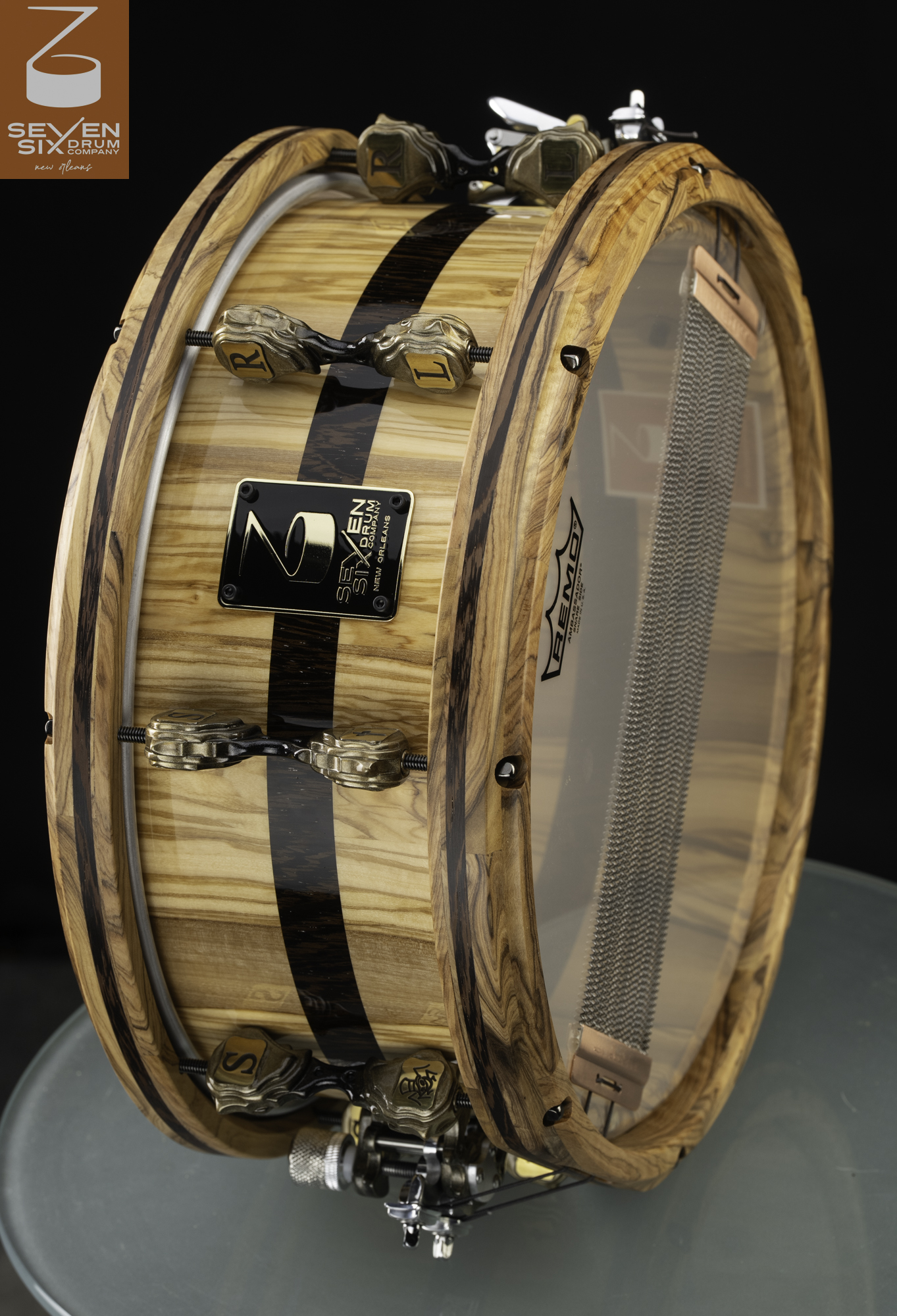 Glossy natural wood snare drum with a dark center stripe, matching wood hoops, and sculpted bronze letter lugs.