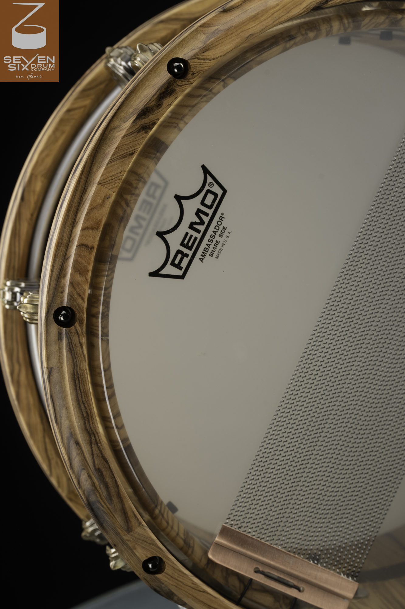 Snare drum bottom featuring gloss-finished figured wood hoops, recessed black tension bolts, and wire snares with a copper clip.