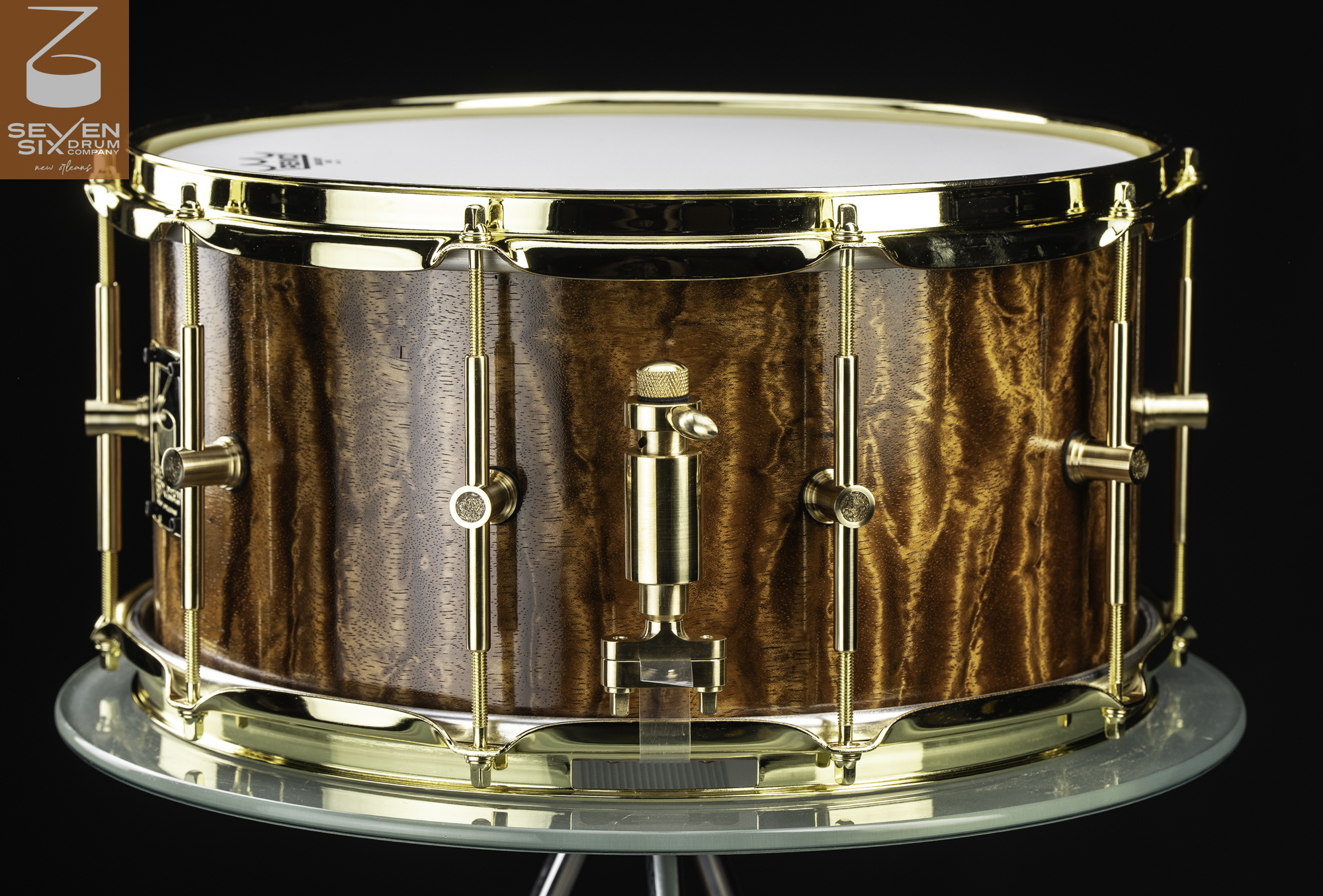 Handmade snare drum featuring a glossy figured wood shell with polished brass hoops, tube lugs, and a brass throw-off.