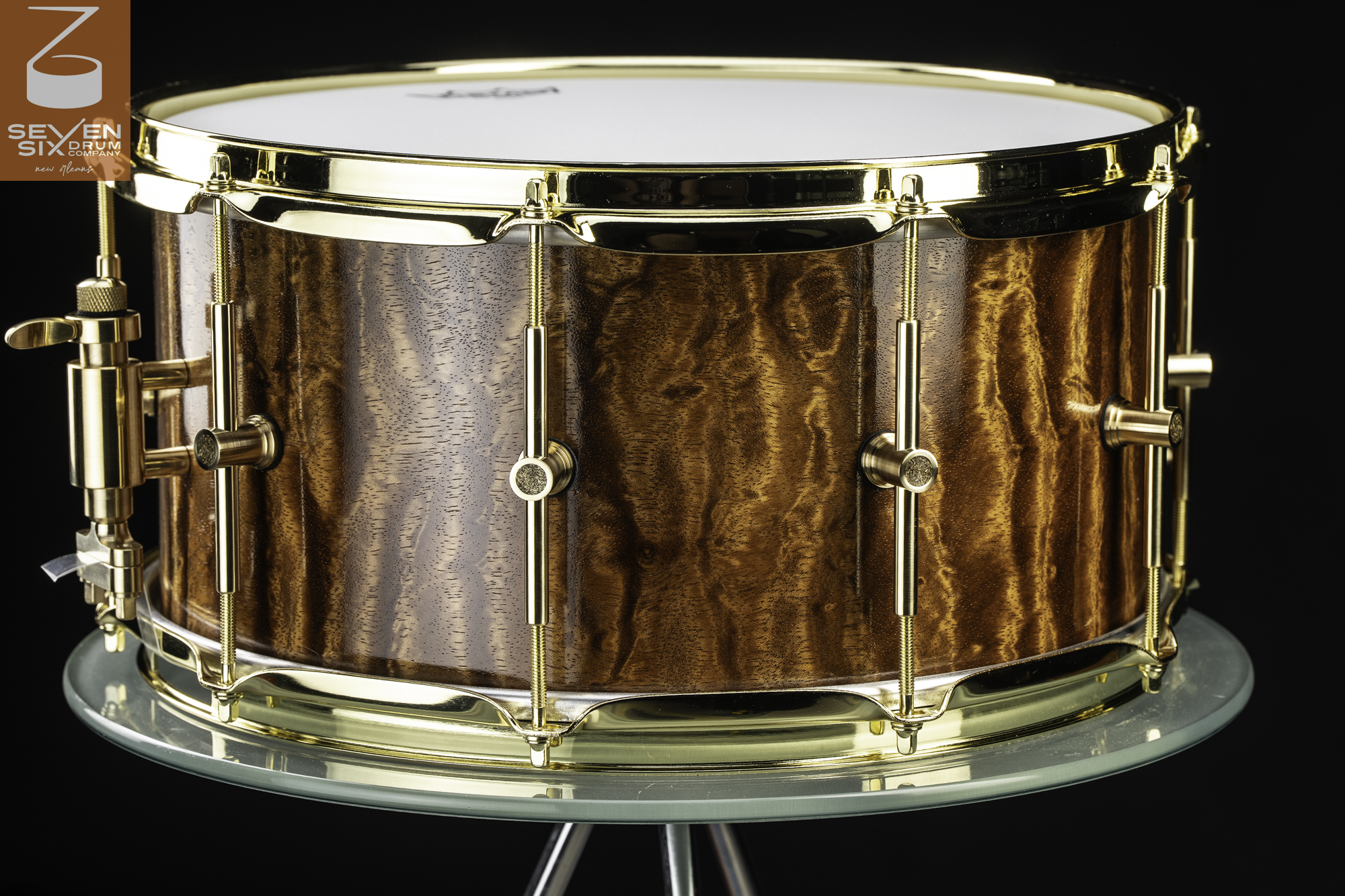Handmade snare drum featuring a glossy figured brown wood shell with polished brass hoops, lugs, and throw-off.