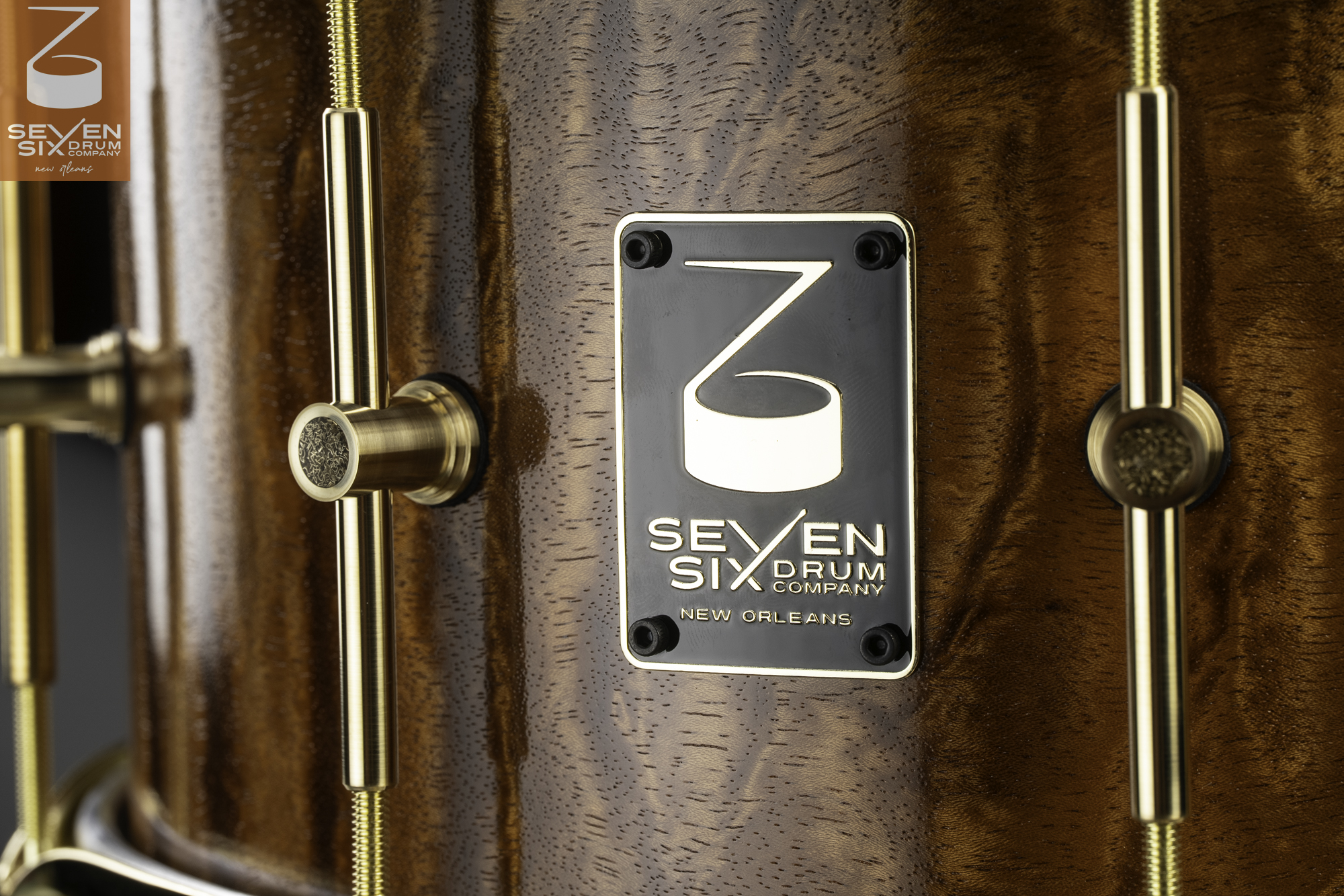 Glossy figured wood snare drum shell featuring brass tube lugs and a black metal logo badge.