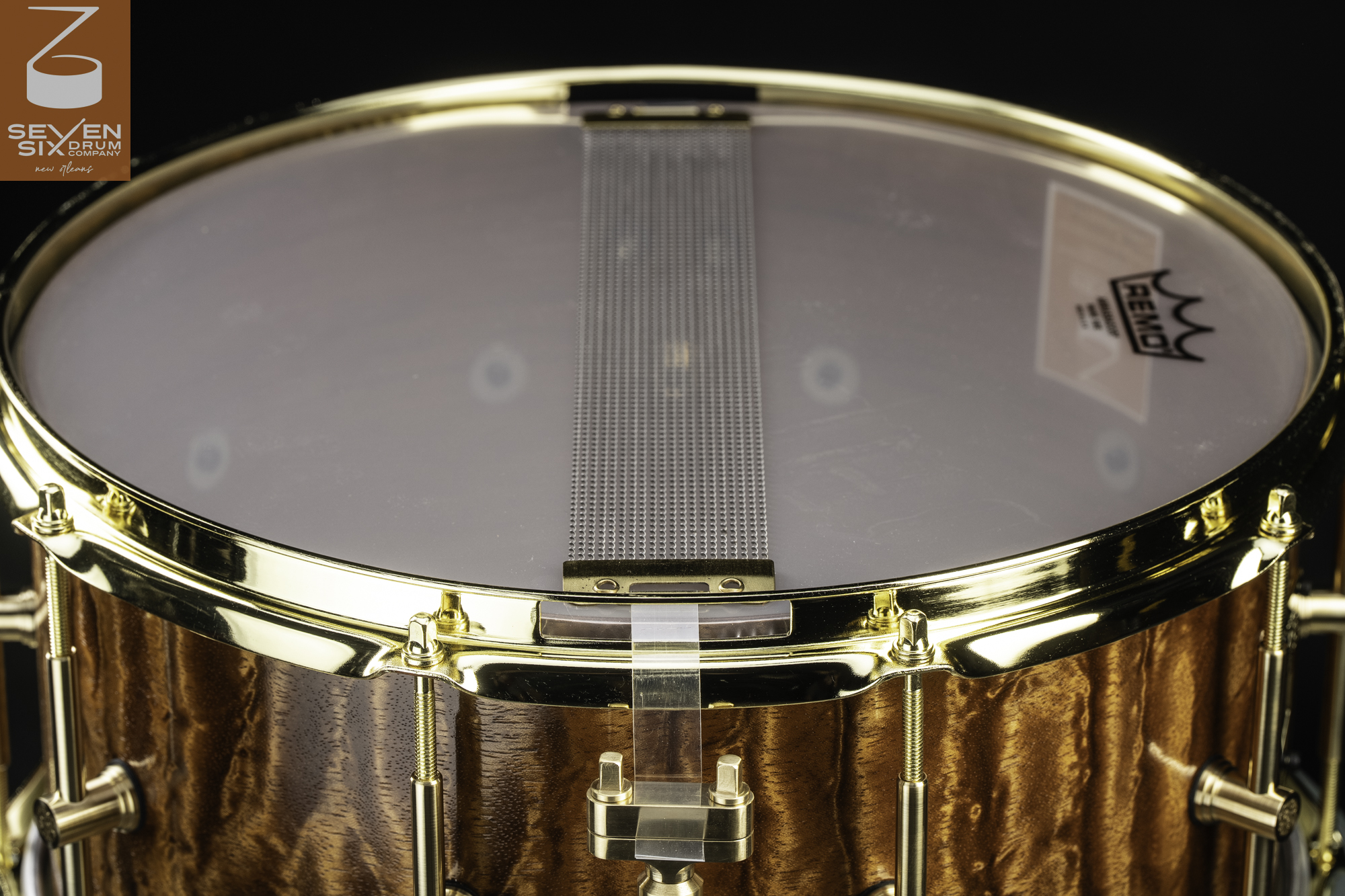Bottom view of a snare drum with a glossy figured wood shell, polished brass hoop, and snare wires stretched across the head.
