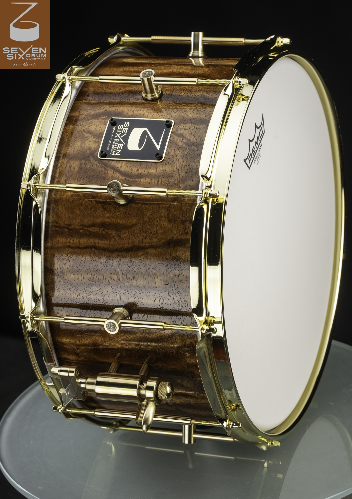 Handmade snare drum with a glossy figured brown wood shell, polished gold hoops, tube lugs, and throw-off.