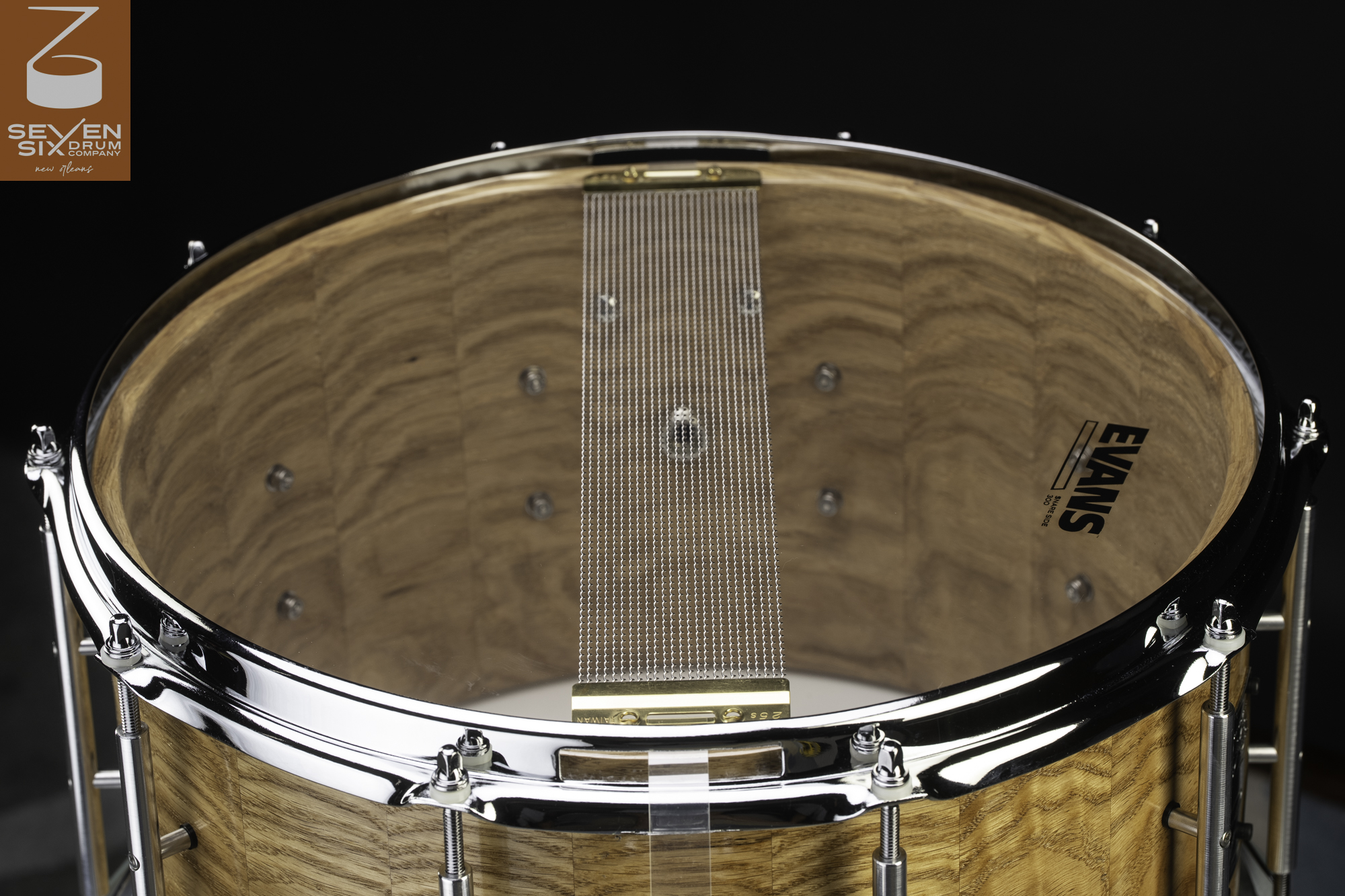 Resonant side of a natural-finish segmented wood snare drum, showing snare wires, clear head, and polished chrome hardware.