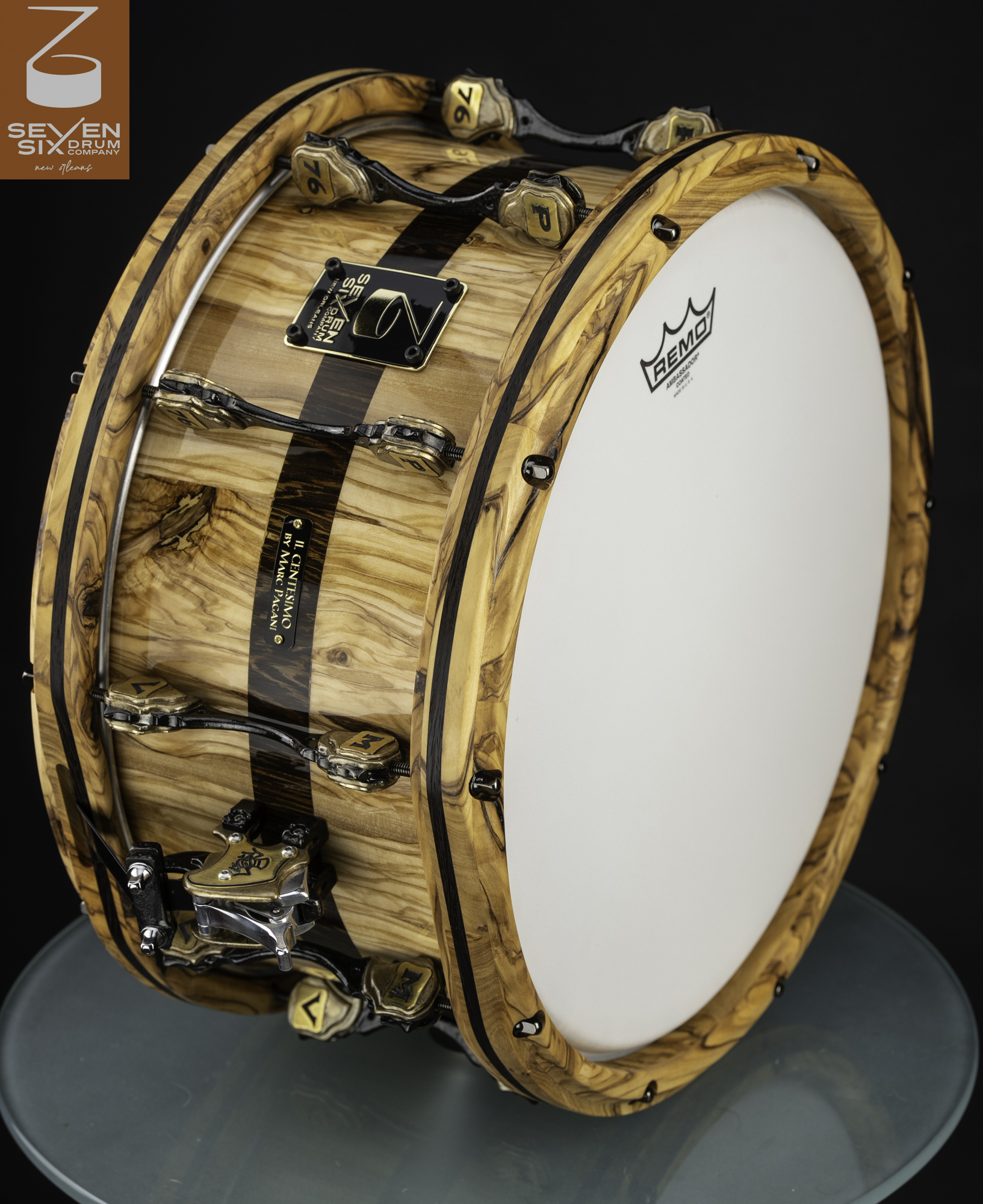 Glossy olive wood snare drum with a dark inlay stripe, matching wooden hoops, and custom black and brass hardware.