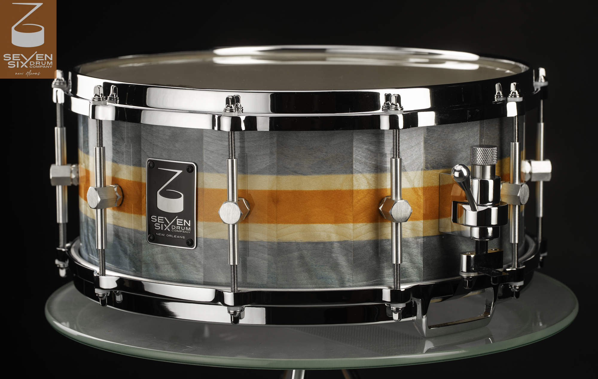Handmade snare drum with a glossy gray and orange striped wood shell, polished chrome hoops, and side throw-off.
